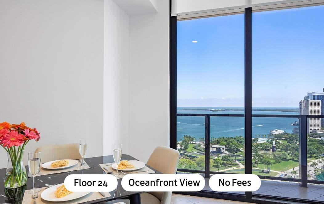 Modern dining area with a black marble-top table set for two, featuring a large floor-to-ceiling window offering panoramic ocean and city views from a high floor.