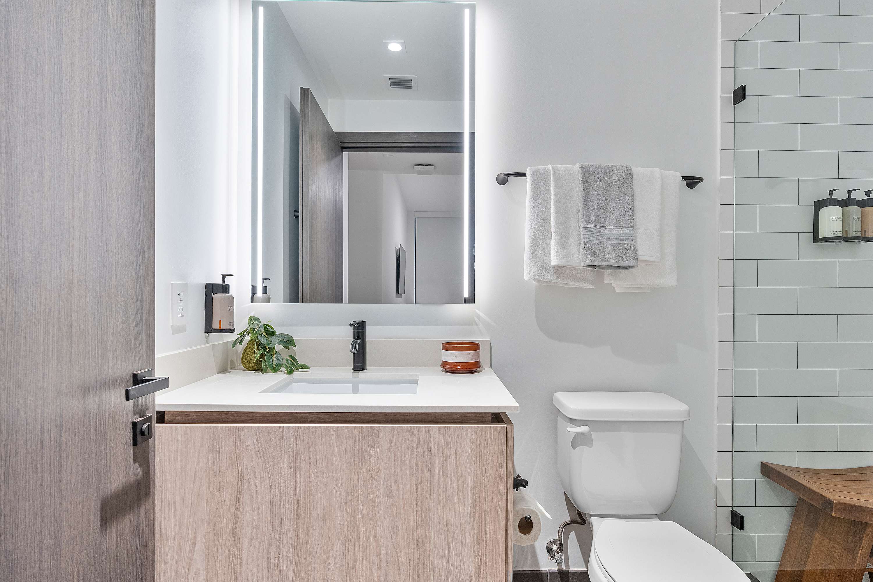 Modern bathroom with a light wood vanity, white countertop, illuminated mirror, and a walk-in shower with white subway tiles and a glass enclosure, featuring a wooden stool inside.