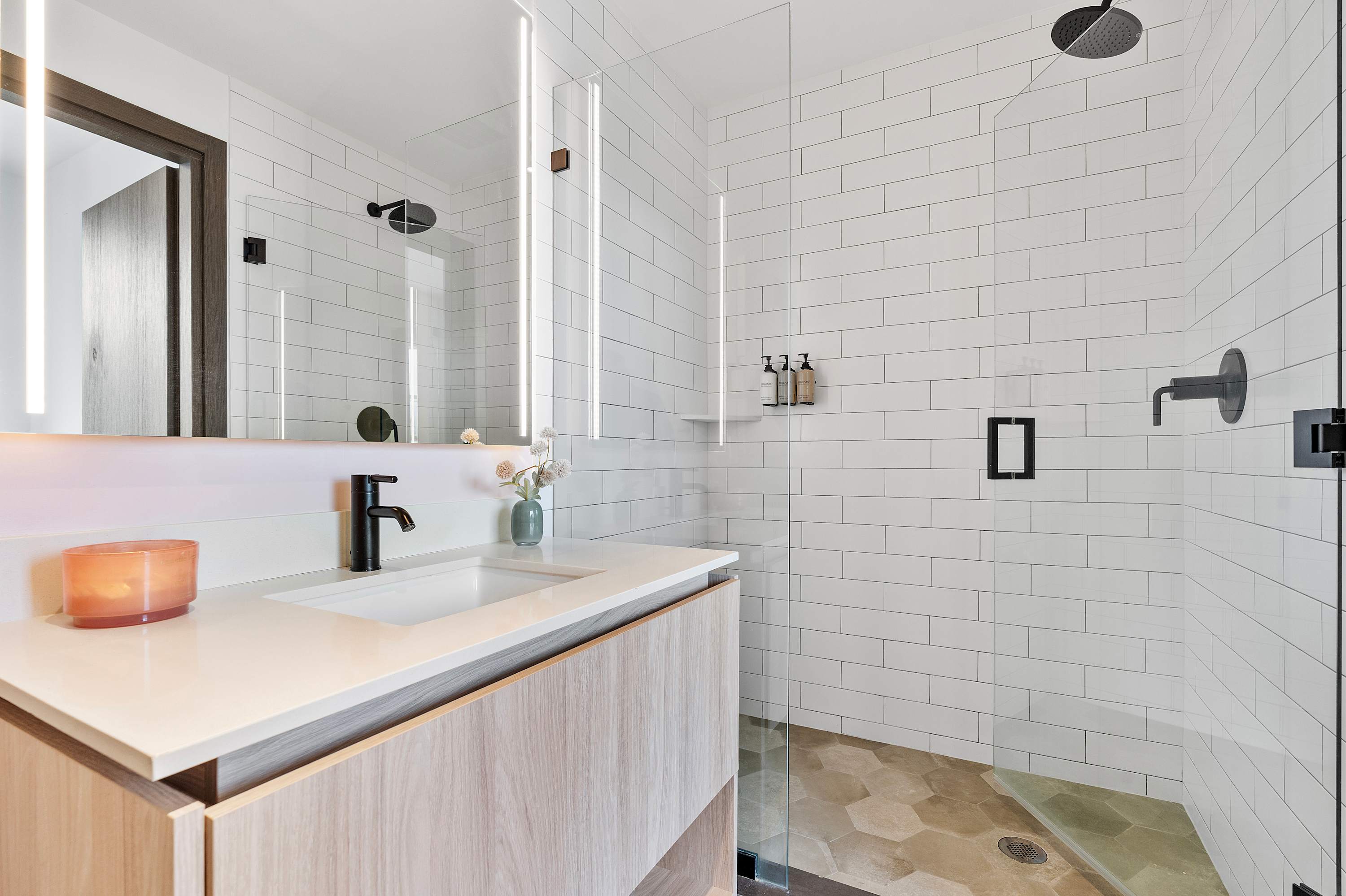 A sleek bathroom featuring a light wood vanity with a white sink and black faucet, an illuminated mirror, and a spacious walk-in shower with white subway tiles and a glass door.
