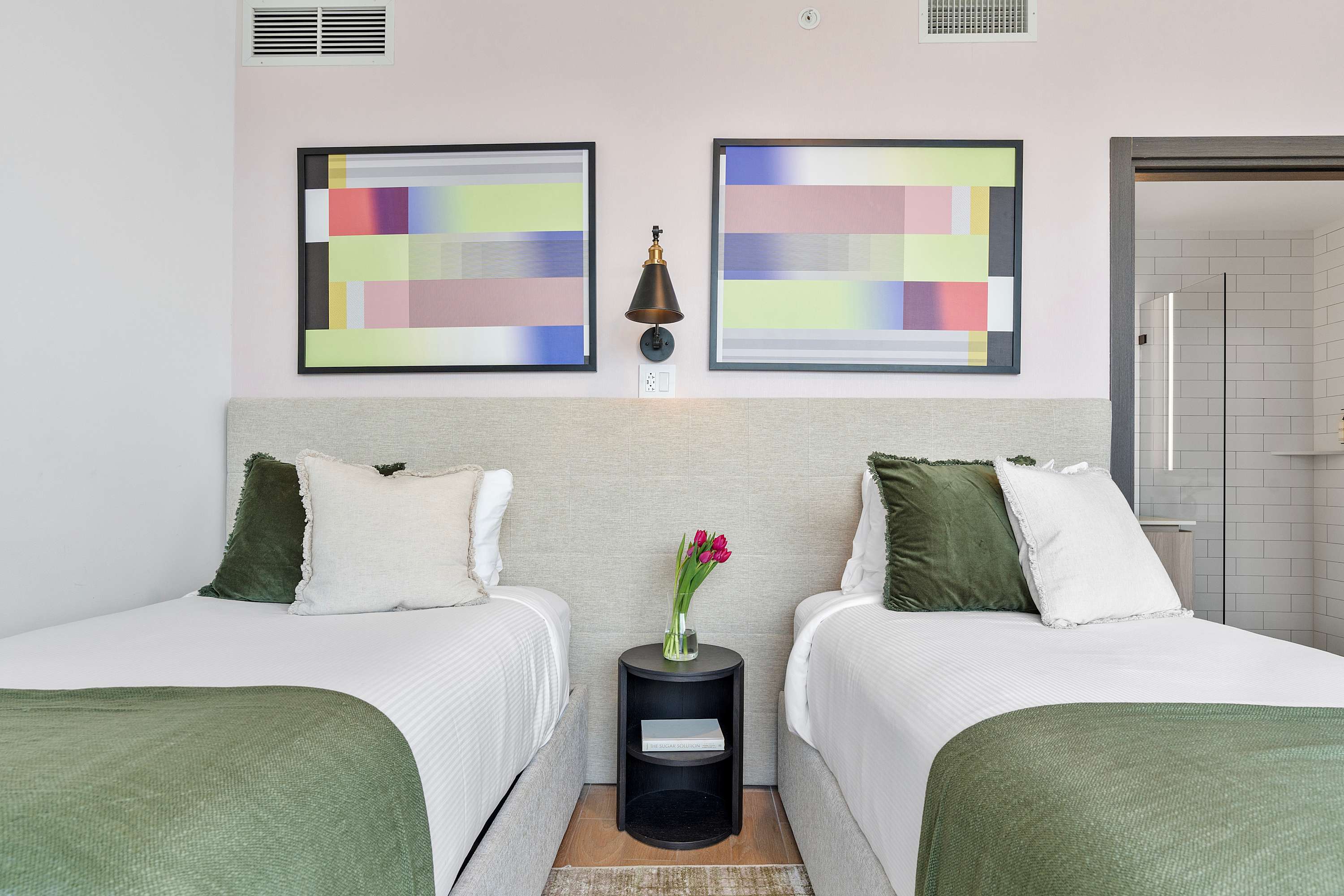 A close-up of two twin beds with light grey headboards, white bedding, and green throws. A small black nightstand with a vase of flowers sits between them, beneath colorful abstract artwork and wall sconces.