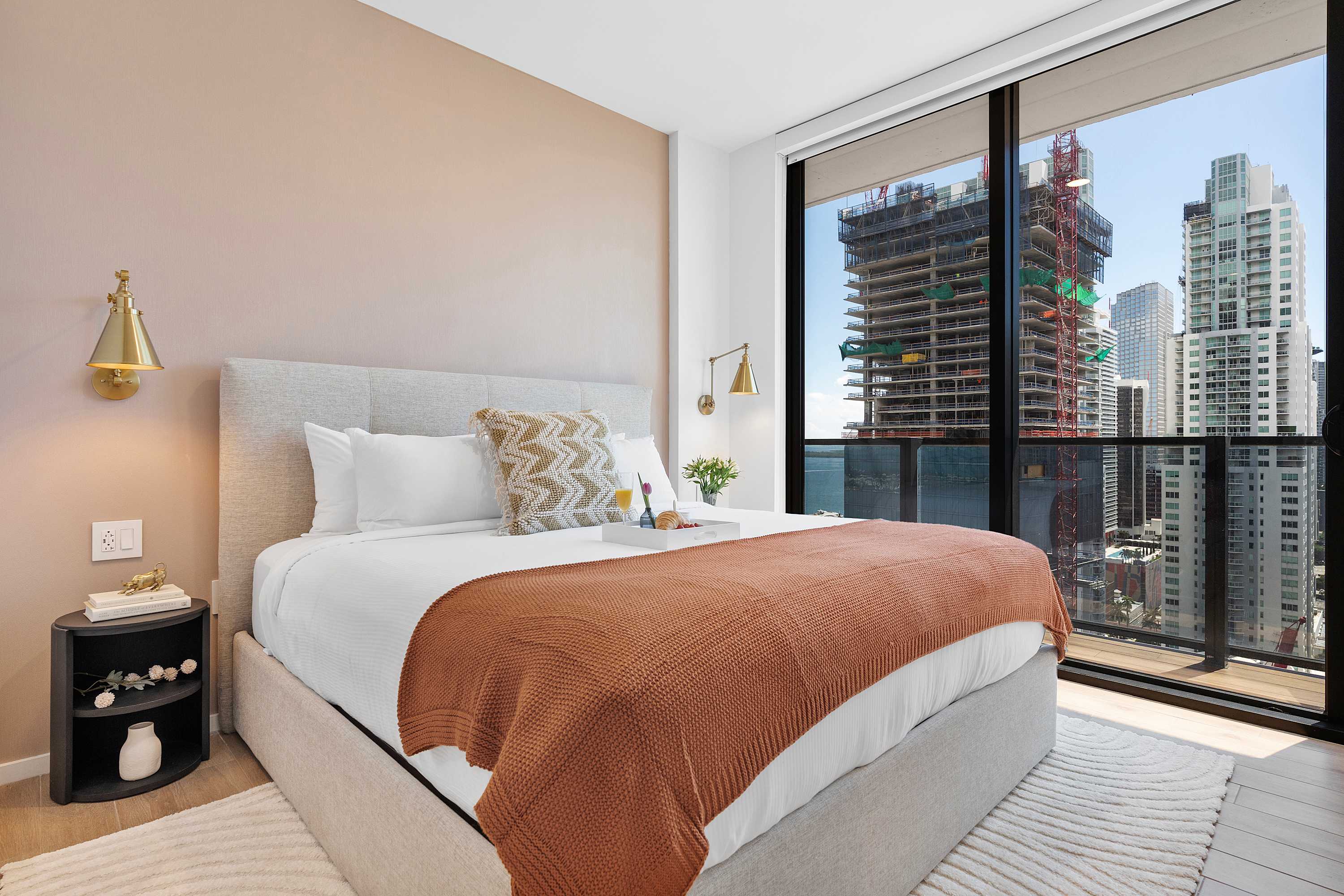 Bright bedroom-1 featuring a queen-sized bed with a light grey upholstered headboard, an orange throw, and gold wall sconces, positioned against a two-tone wall and facing floor-to-ceiling windows with city views.