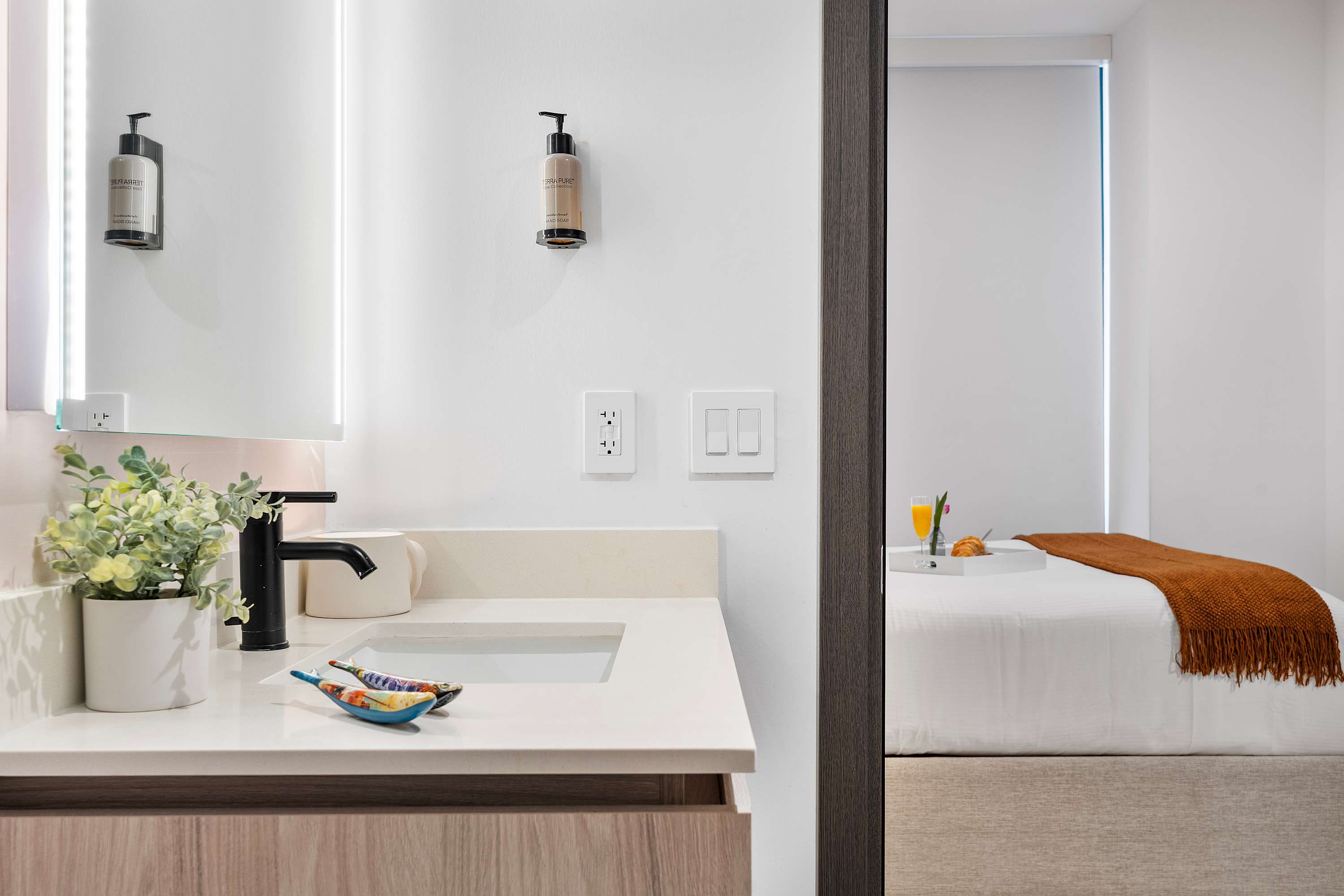A modern bathroom vanity with a light wood cabinet, white countertop, black faucet, and an illuminated mirror, offering a glimpse into the adjacent bedroom with a bed and breakfast tray.