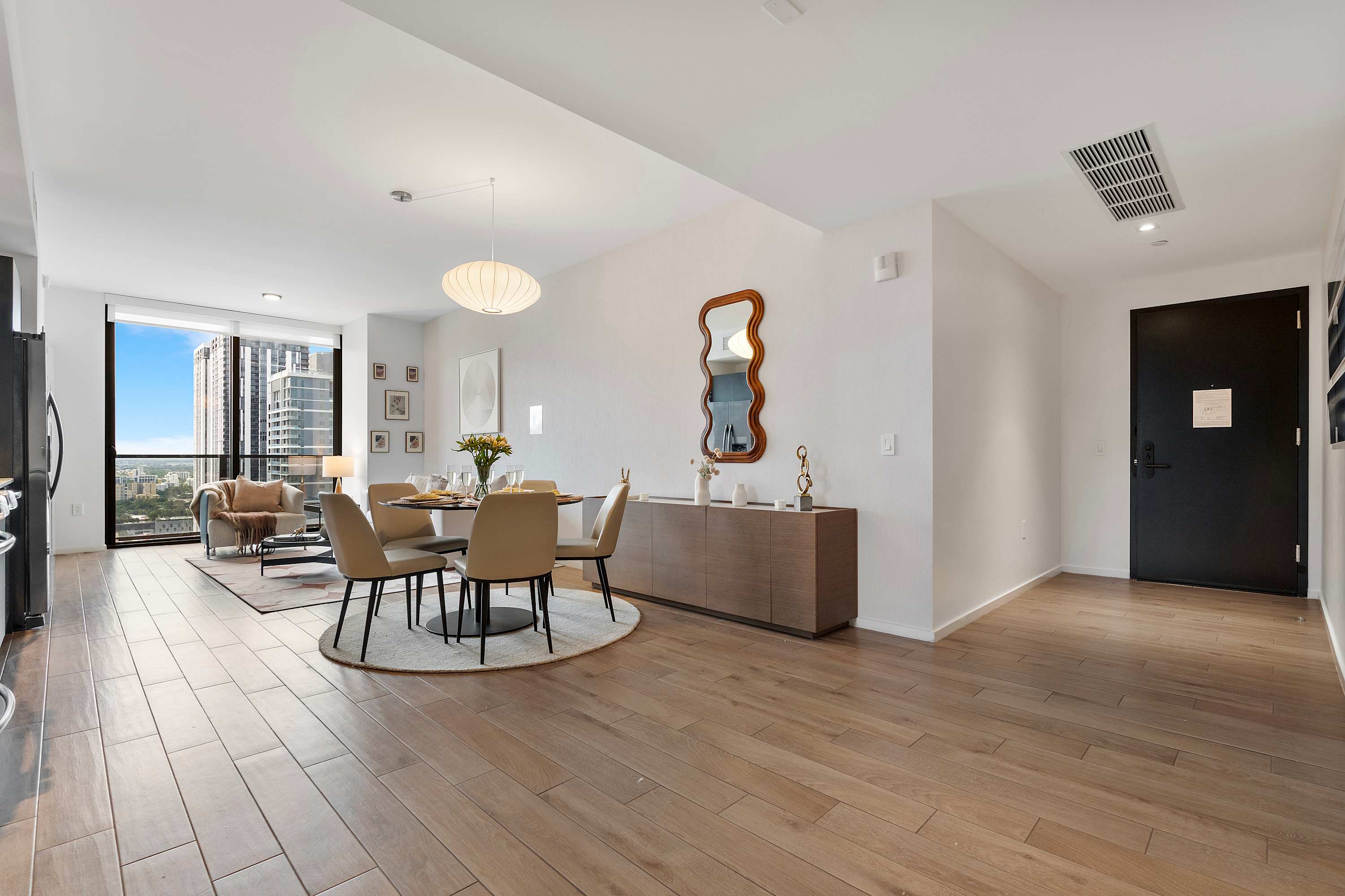 Open-concept dining area with a round black table and four beige chairs, illuminated by a modern pendant light, adjacent to a wooden credenza with a decorative mirror, and leading into the living room and entryway.