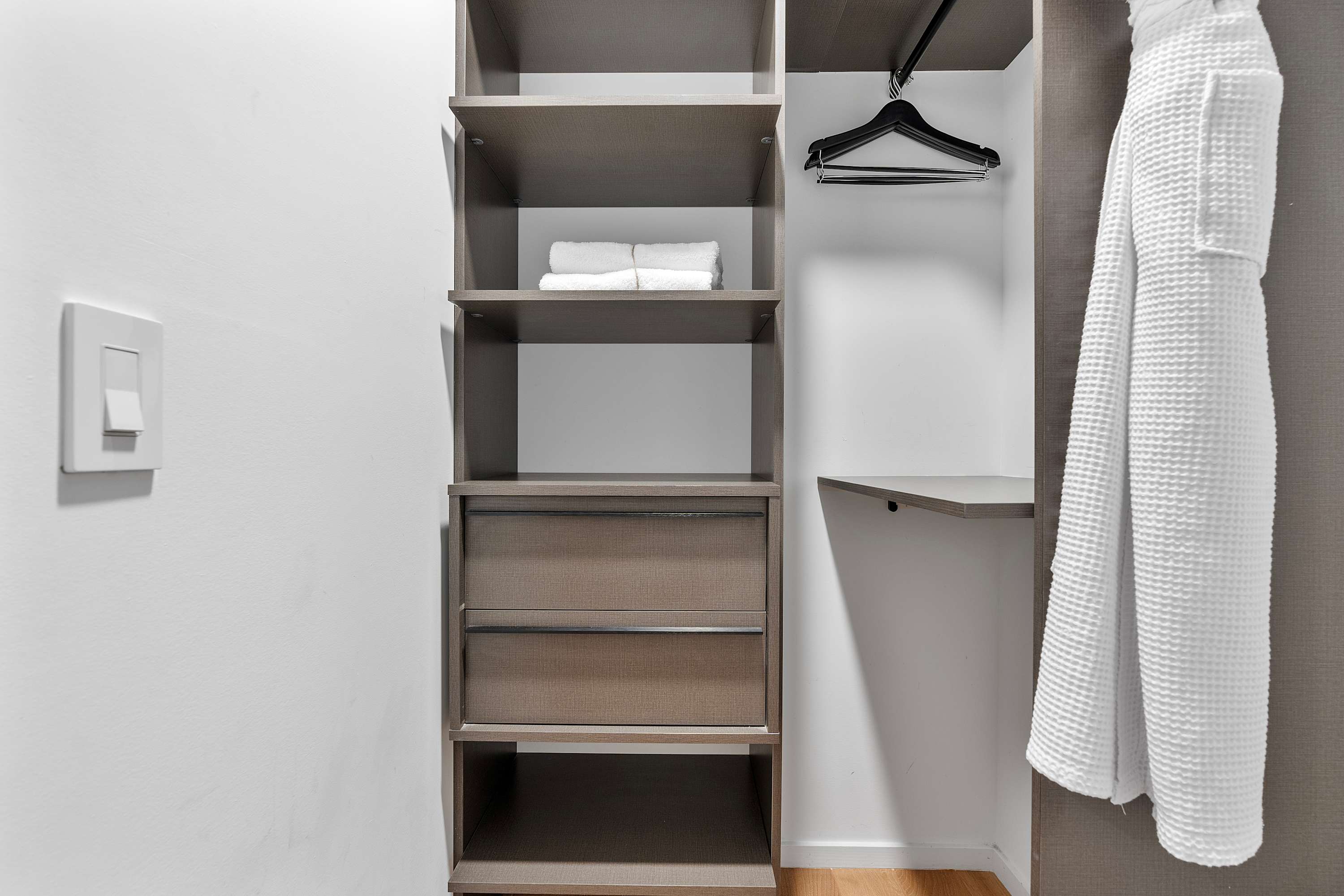 A well-organized closet with built-in shelving, drawers, and hanging space, featuring folded white towels and a white bathrobe hanging on a rod.