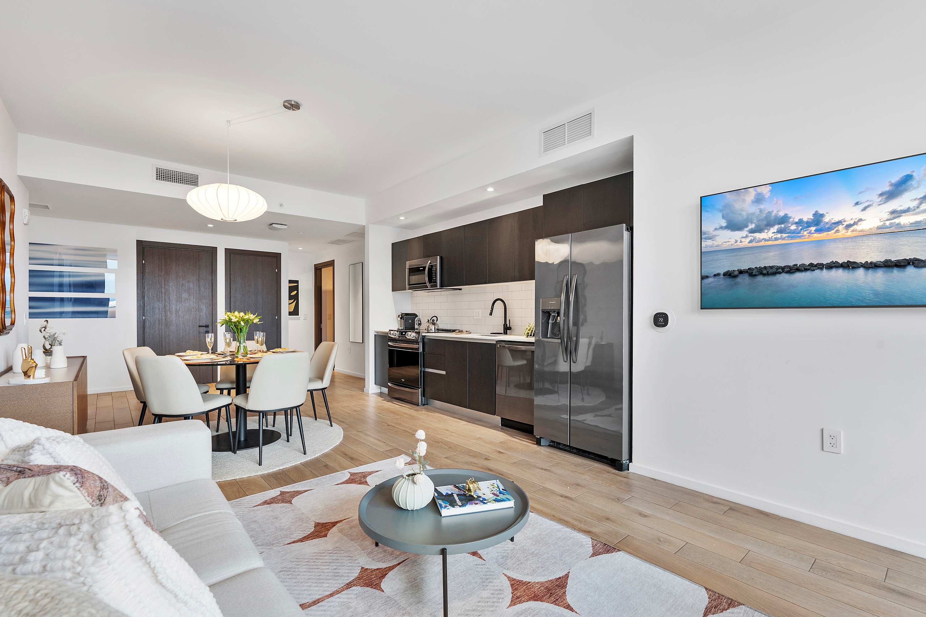 Wide shot of an open-concept living area and kitchen, featuring a light grey sofa, a round coffee table, and a dark-cabinet kitchen with stainless steel appliances. The space is unified by light hardwood floors and a wall-mounted TV.