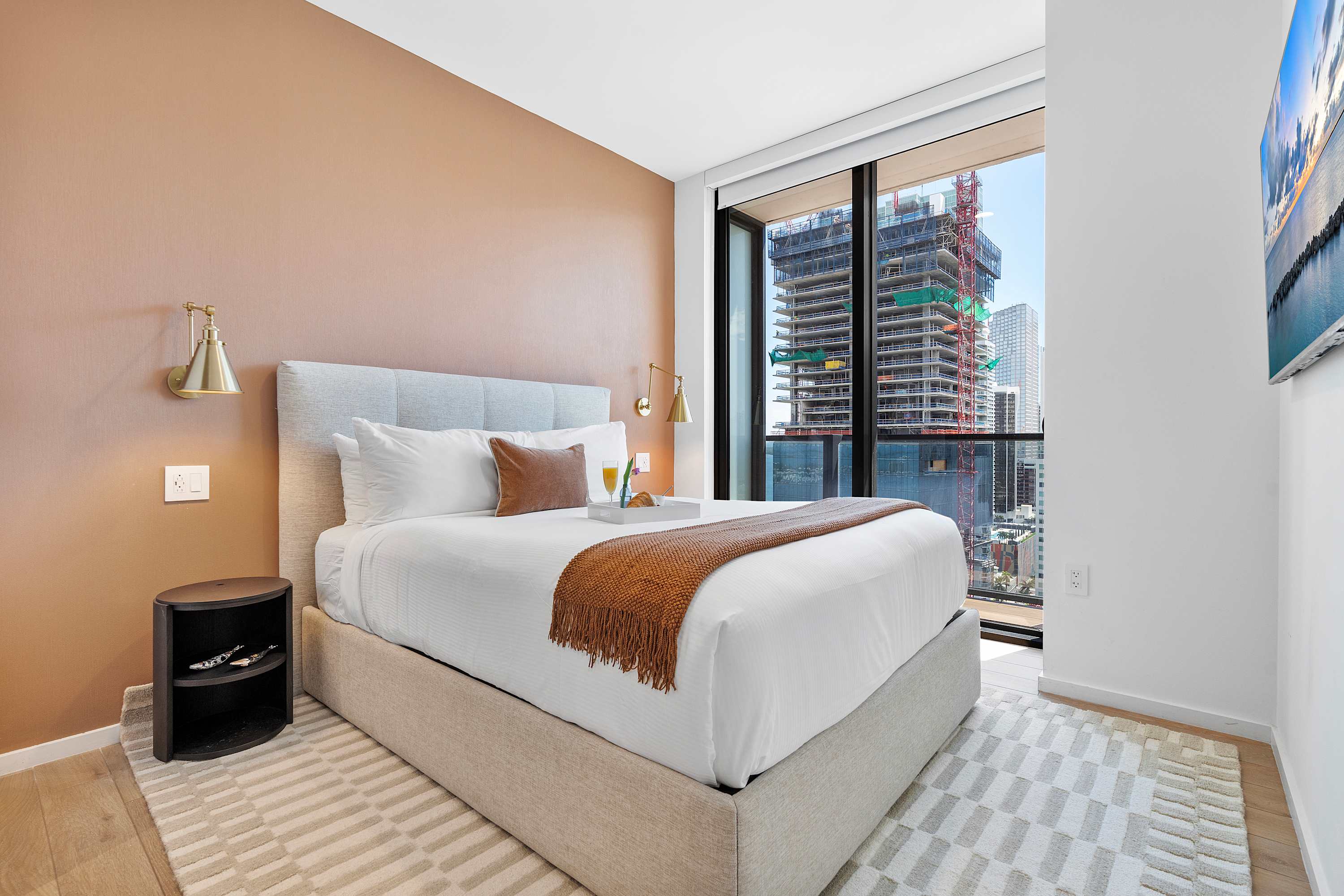 A contemporary bedroom with a large bed, a brown accent wall, and floor-to-ceiling windows offering a city view with construction. A small nightstand and wall sconces are visible.