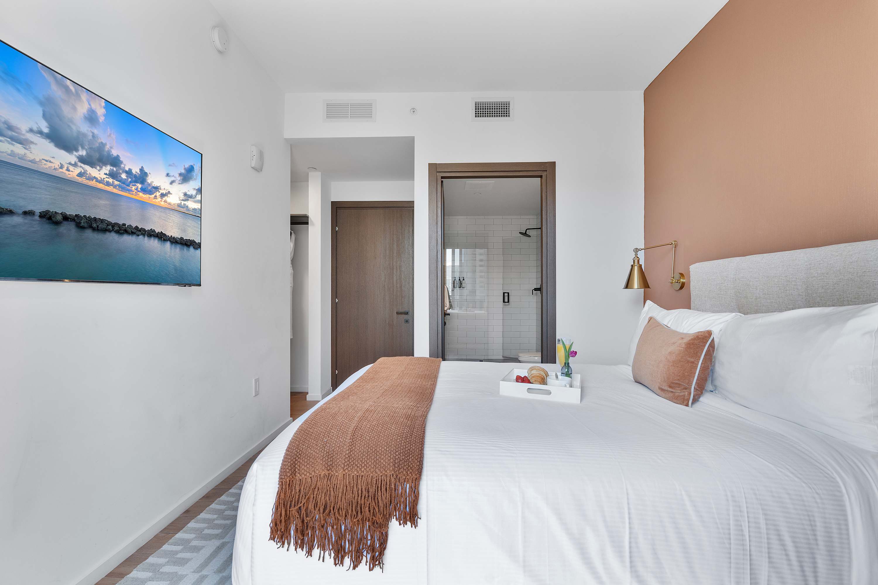 A modern bedroom featuring a large bed with white bedding and a brown throw, a wall-mounted TV displaying a seascape, and a partial view into a sleek bathroom with a glass shower.
