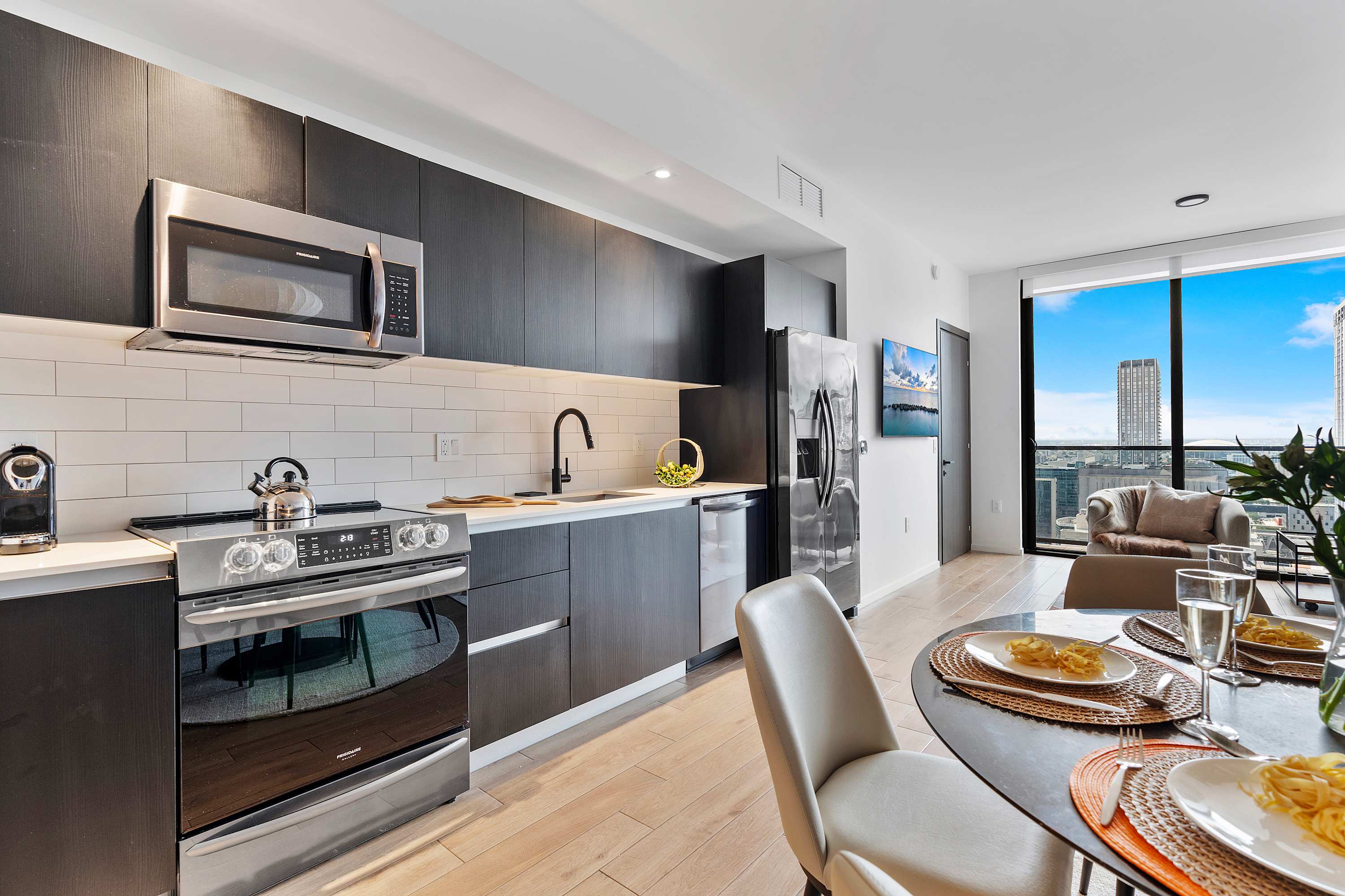Modern kitchen featuring dark flat-panel cabinetry, stainless steel appliances including a microwave, oven, and refrigerator. A white subway tile backsplash completes the look, with light hardwood floors extending into the open living area.
