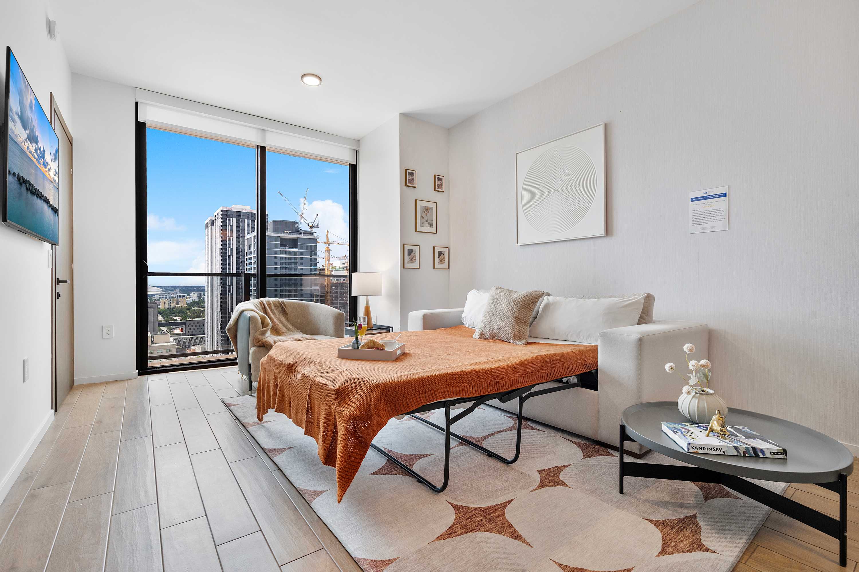 Bright living room with a sofa bed pulled out, covered with an orange throw, facing a large window with city views. A small round coffee table sits on a patterned rug, and a wall-mounted TV is visible on the right.