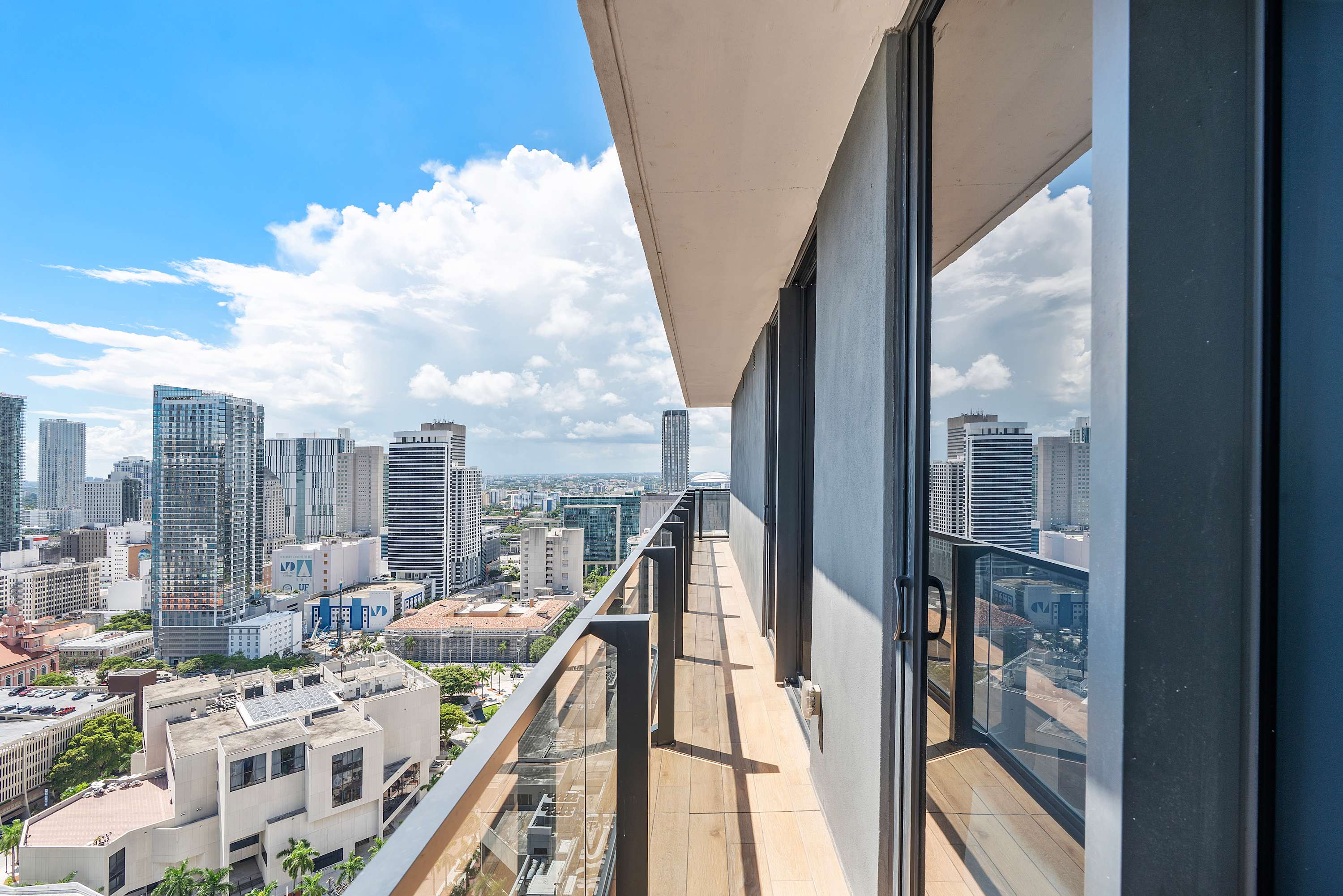 Long, narrow balcony with glass railings offering panoramic city views of multiple high-rise buildings under a blue sky with white clouds. The floor is light wood-look tile.