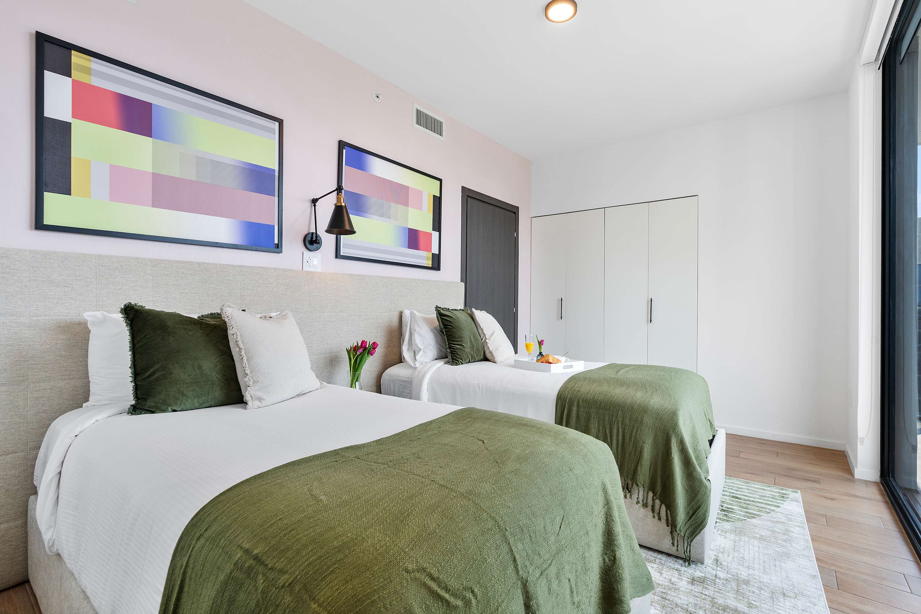 A spacious bedroom with two twin beds, white bedding, and green throws, positioned on a light-colored area rug. The room features colorful abstract art, a large window, and a white built-in closet.