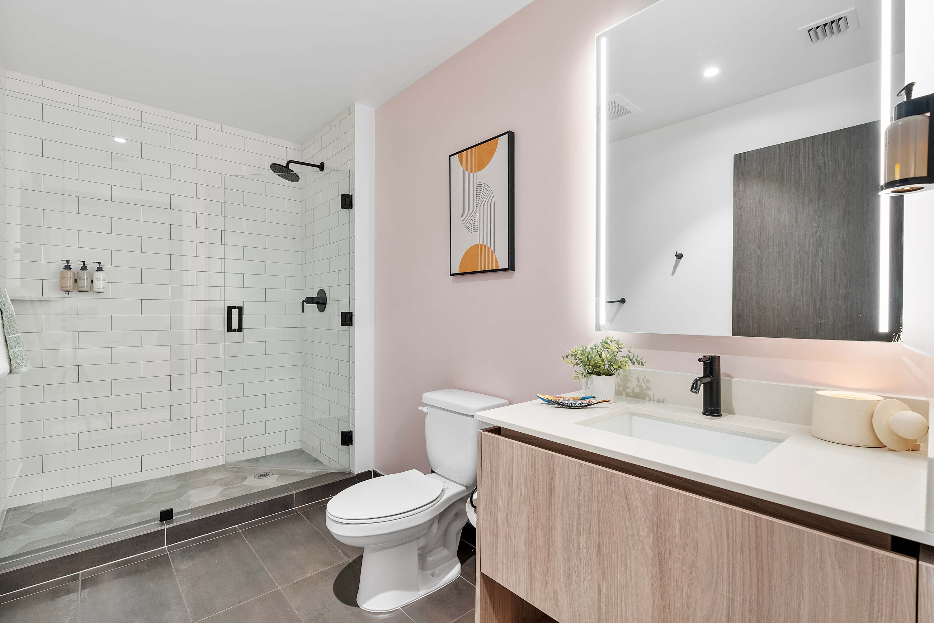 A contemporary bathroom featuring a white toilet, a light wood vanity with a white countertop, and a spacious walk-in shower with white subway tiles and a glass enclosure. A pink accent wall adds a touch of color.