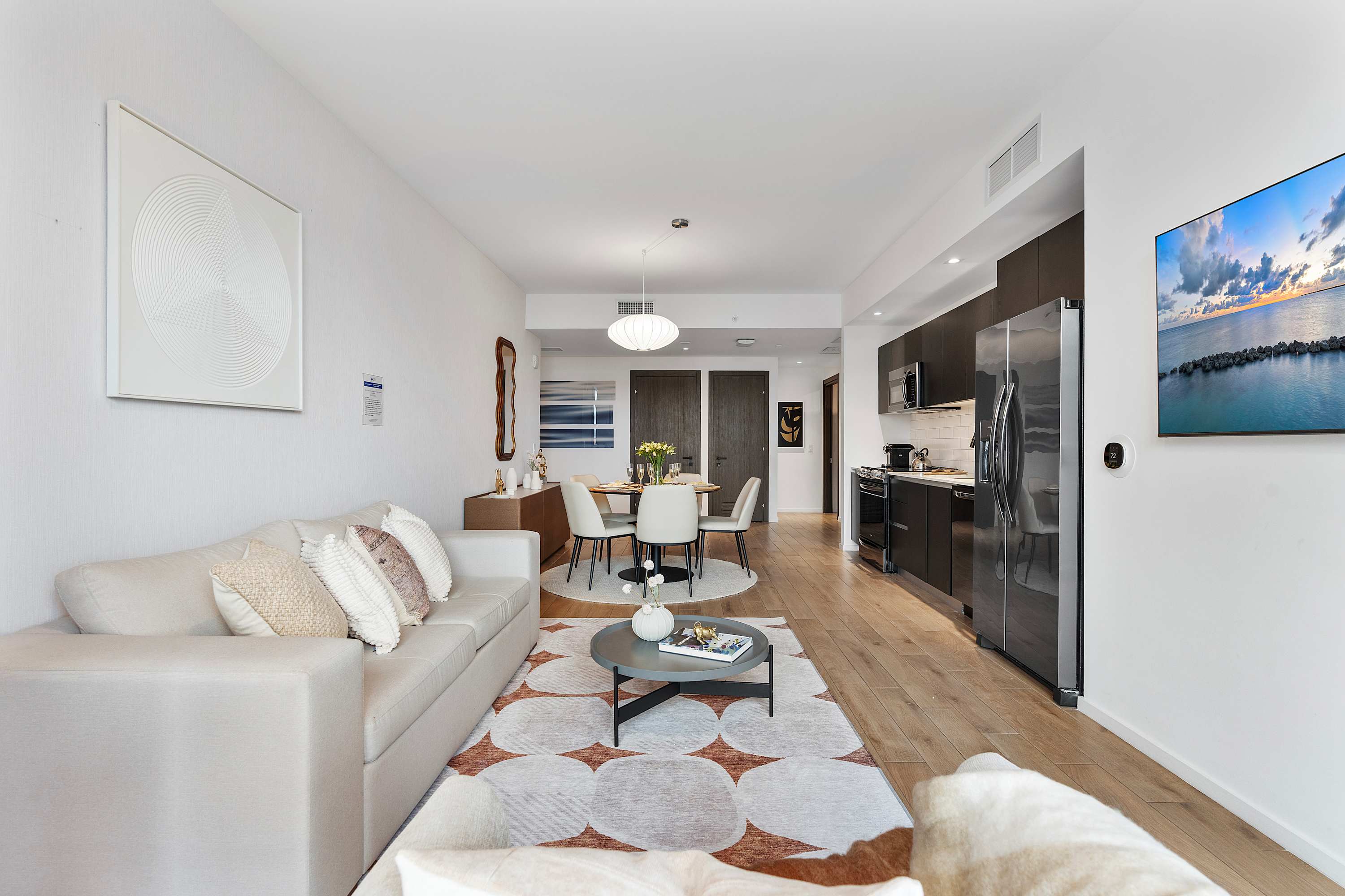 Modern open-concept living space featuring a light grey sofa with decorative pillows, a round coffee table on a patterned rug, and a dining area with a round table and four chairs, all leading into a sleek kitchen with dark cabinetry.