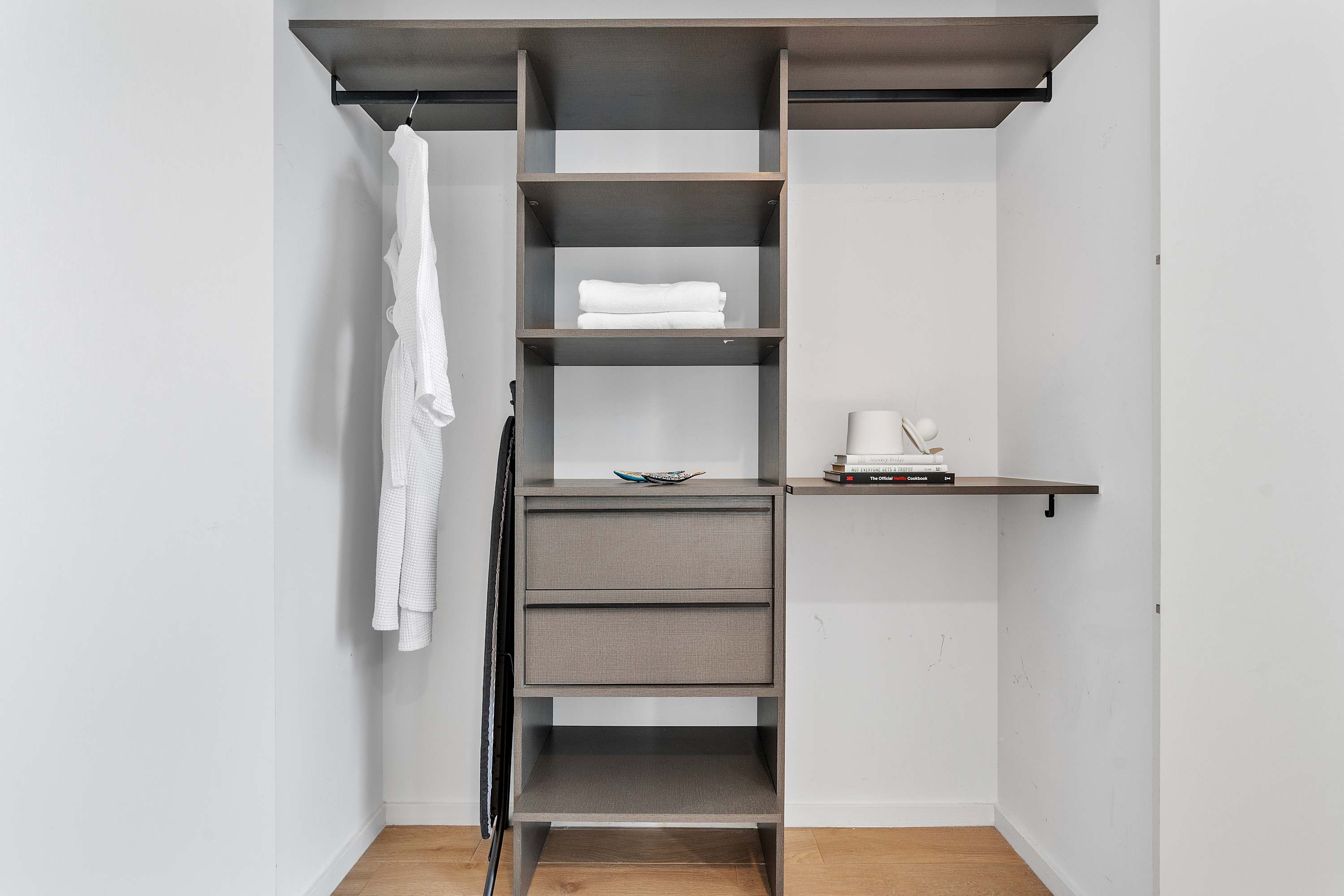 A modern, built-in closet system features dark wood shelves, two drawers, and a hanging rod with a white robe, all set against white walls and light wood flooring.