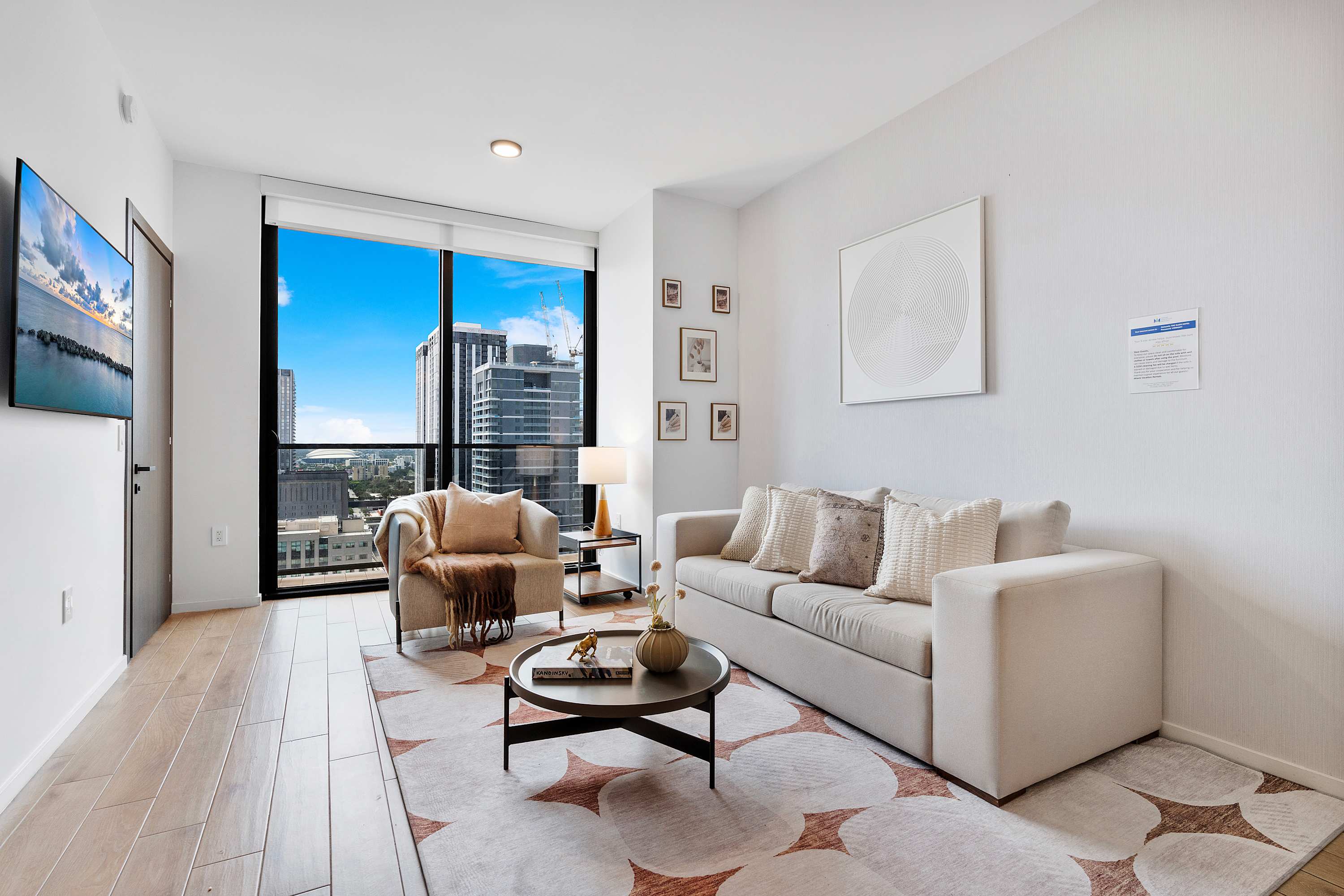 Spacious living room with a light grey sofa, an armchair, and a round coffee table on a patterned rug. The room features a large window with a city view and minimalist wall art, complemented by light hardwood floors.