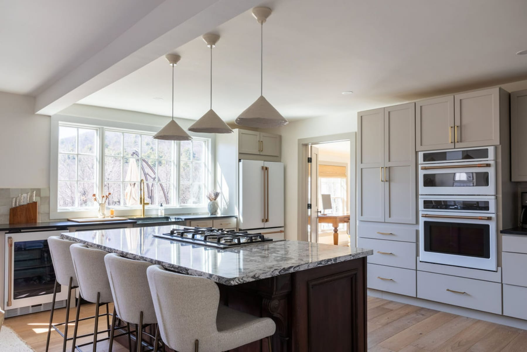 Bright kitchen with a large island featuring a gas cooktop and bar seating, light gray cabinetry with integrated ovens, and pendant lights hanging above the island. A large window provides natural light and views.