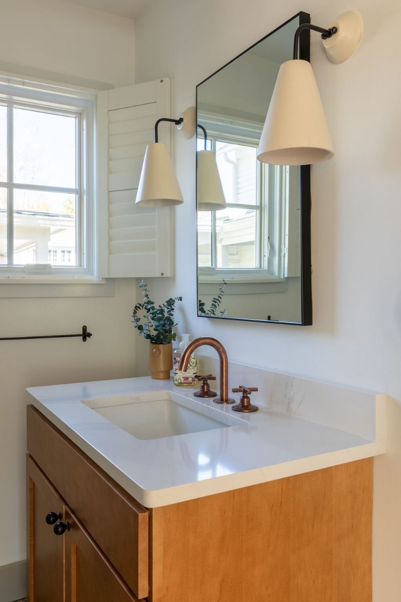A bright bathroom vanity with a light wood cabinet, white quartz countertop, and a modern copper faucet. A rectangular mirror reflects a window with white louvered shutters, complemented by two contemporary wall sconces.