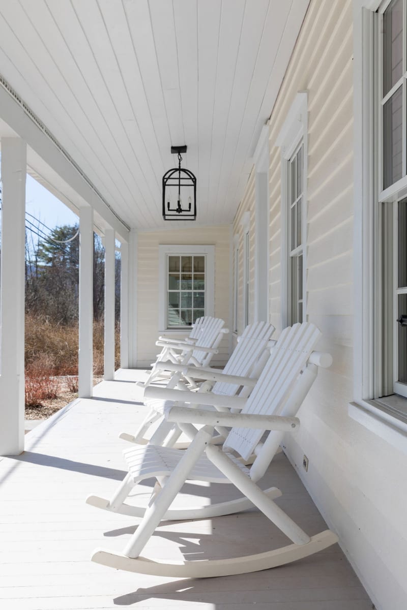 A long, inviting porch is furnished with several white rocking chairs, set against white siding and windows, with a black lantern light fixture overhead and distant trees.