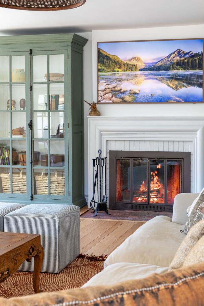 Cozy living room featuring a white fireplace with a burning fire, a large framed landscape art piece above, and a sage green glass-front cabinet next to it. Striped ottomans and a cream sofa are visible in the foreground.