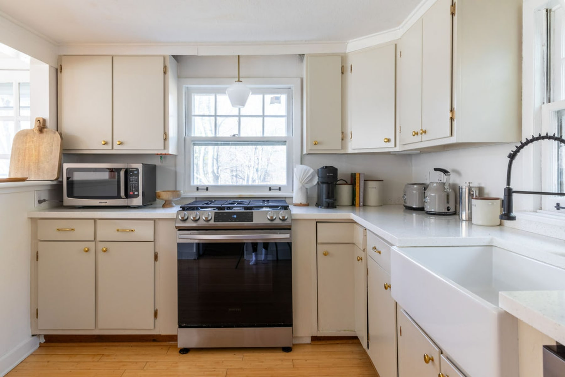 A cozy kitchen featuring light beige cabinets, white countertops, and stainless steel appliances including a range and microwave. A white farmhouse sink is positioned under a window, providing natural light to the space.