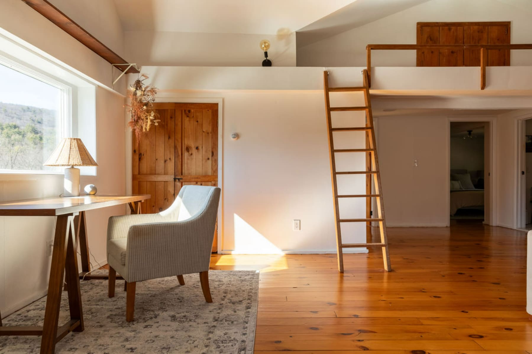 Bright room with polished wooden floors, a desk by a large window, and a striped armchair. A wooden ladder leads to a loft space above, with a rustic wooden door visible in the background.