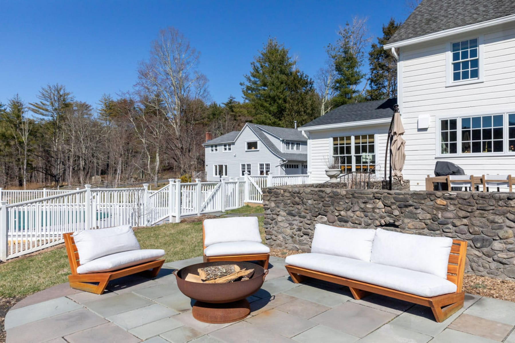 Stone patio with a metal fire pit surrounded by modern wooden outdoor seating with white cushions, set against a backdrop of a white house and distant bare trees under a clear blue sky.