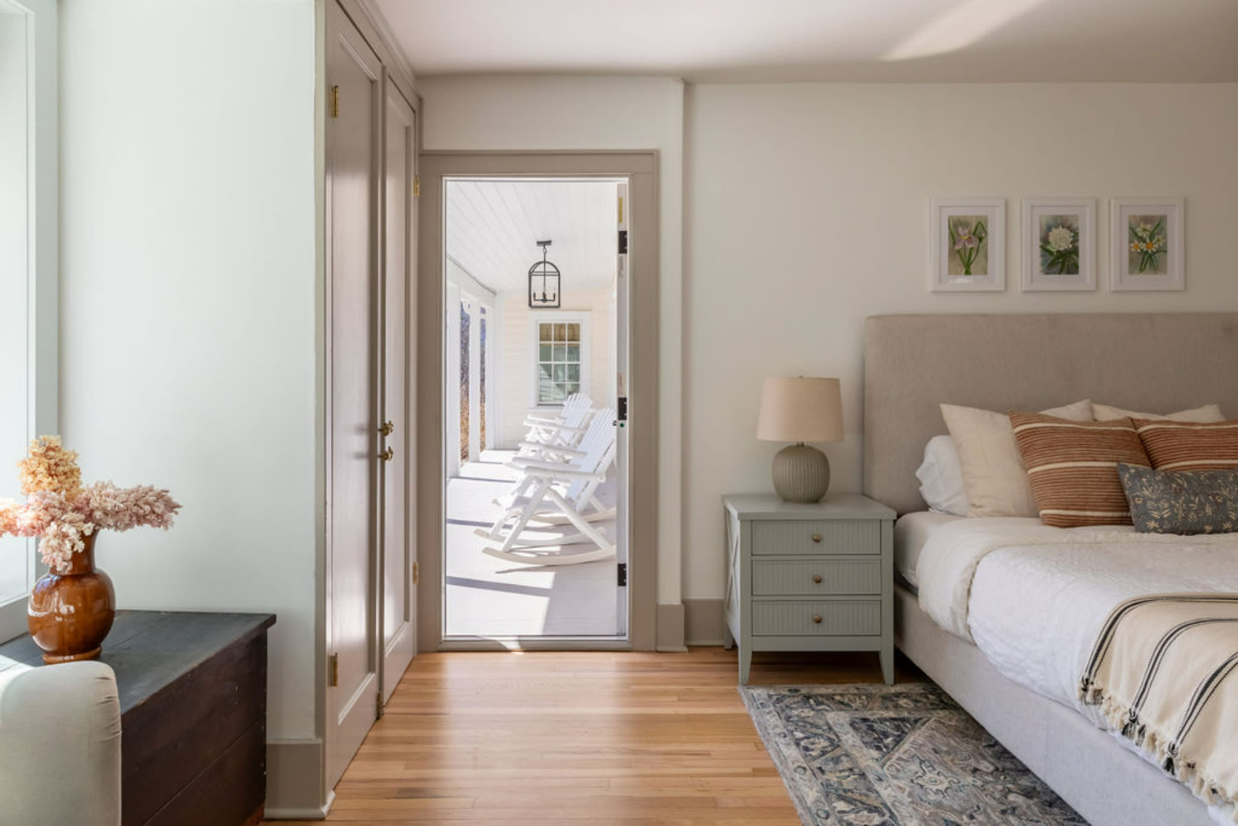 A cozy bedroom features a queen bed with an upholstered headboard, a grey nightstand with a lamp, and hardwood floors, with a door opening to a porch lined with white rocking chairs.
