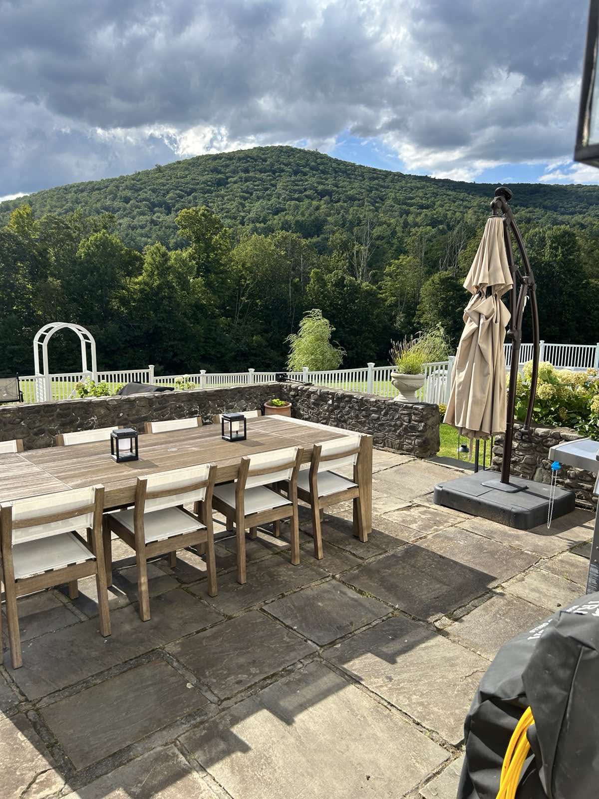 Expansive stone patio featuring a large wooden outdoor dining table with chairs, shaded by an umbrella, overlooking a lush green mountain and white fenced garden under a dramatic cloudy sky.
