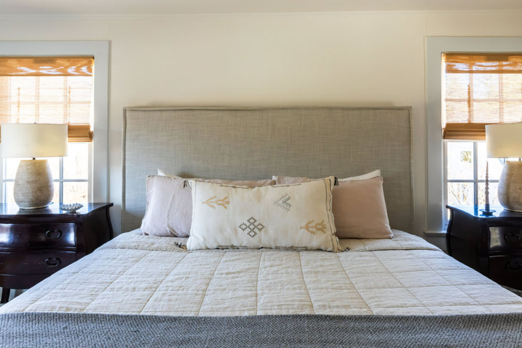 A cozy bedroom centered around a large bed with a light gray upholstered headboard and neutral quilted bedding. Two dark wood nightstands flank the bed, each with a textured lamp and a window adorned with bamboo blinds.