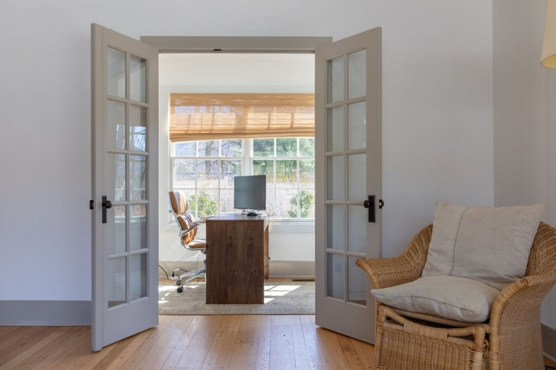 View through open French doors into a bright home office featuring a wooden desk with a computer monitor, a brown leather office chair, and a large window with a woven Roman shade. A wicker armchair is visible in the foreground.