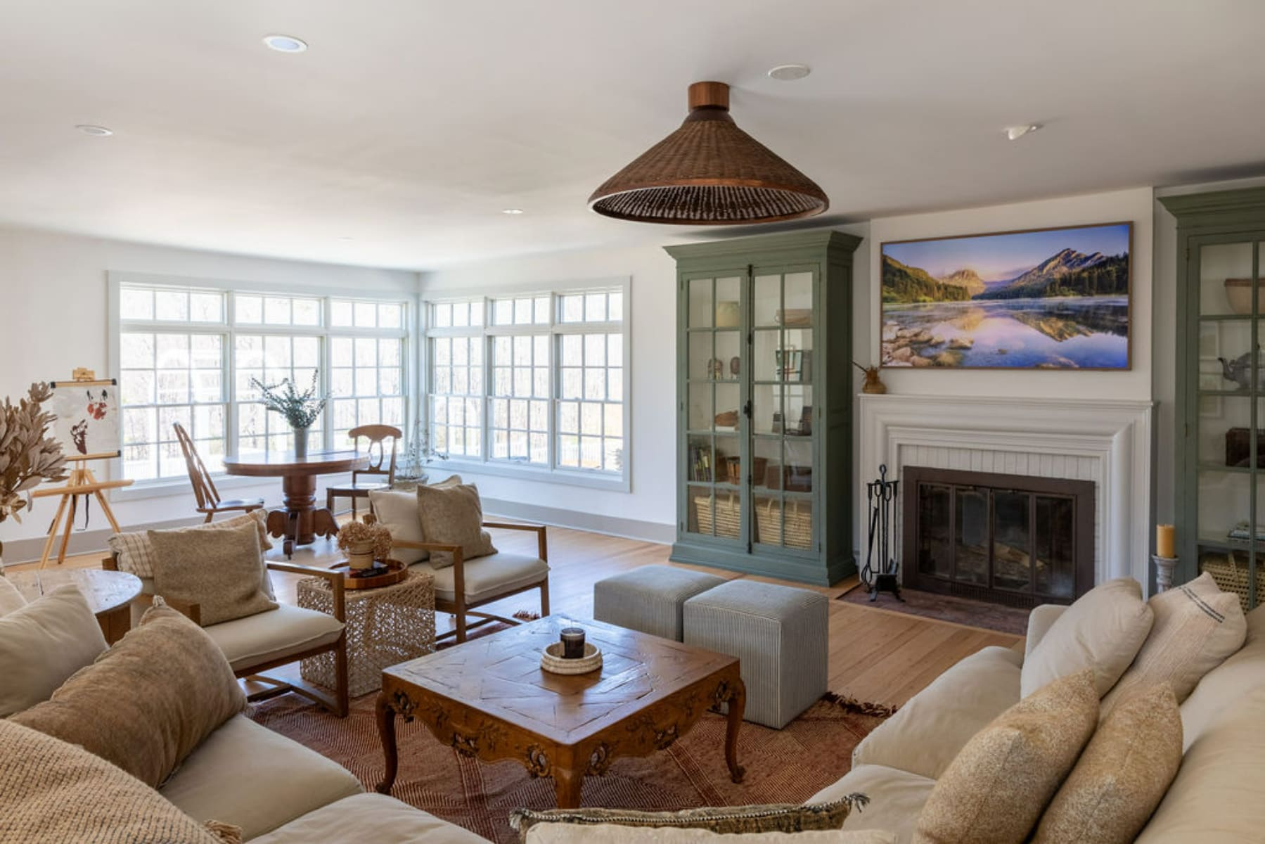 Spacious living room with light hardwood floors, a large bay window overlooking a wooded area, and a central seating arrangement around a carved wooden coffee table. A large woven pendant light hangs from the ceiling.