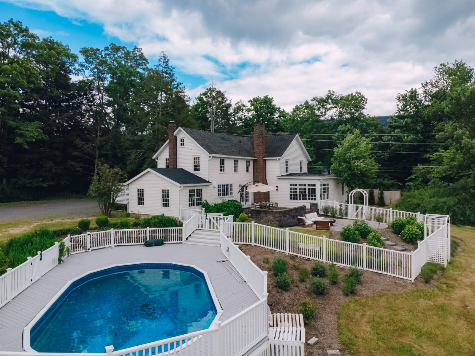 Aerial view of a large white farmhouse with multiple chimneys, surrounded by lush green trees and a manicured lawn. A fenced outdoor pool with a deck is visible in the foreground, with a patio area near the house.