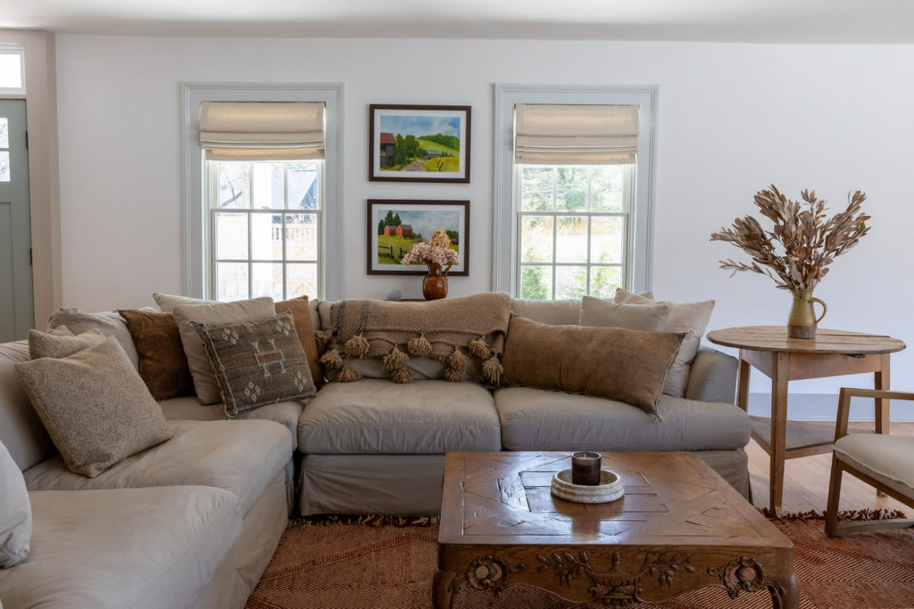 Bright living room corner with a large L-shaped sectional sofa adorned with various throw pillows and a textured blanket. Two windows with roman shades are above a small wooden table with framed artwork.