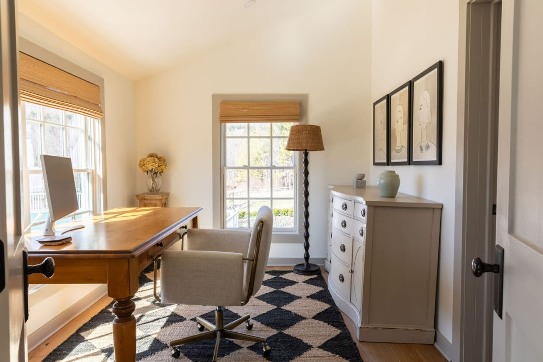 Cozy home office with a wooden desk, a comfortable office chair, and a light gray dresser with framed abstract art above it. A black and white geometric rug covers part of the hardwood floor.