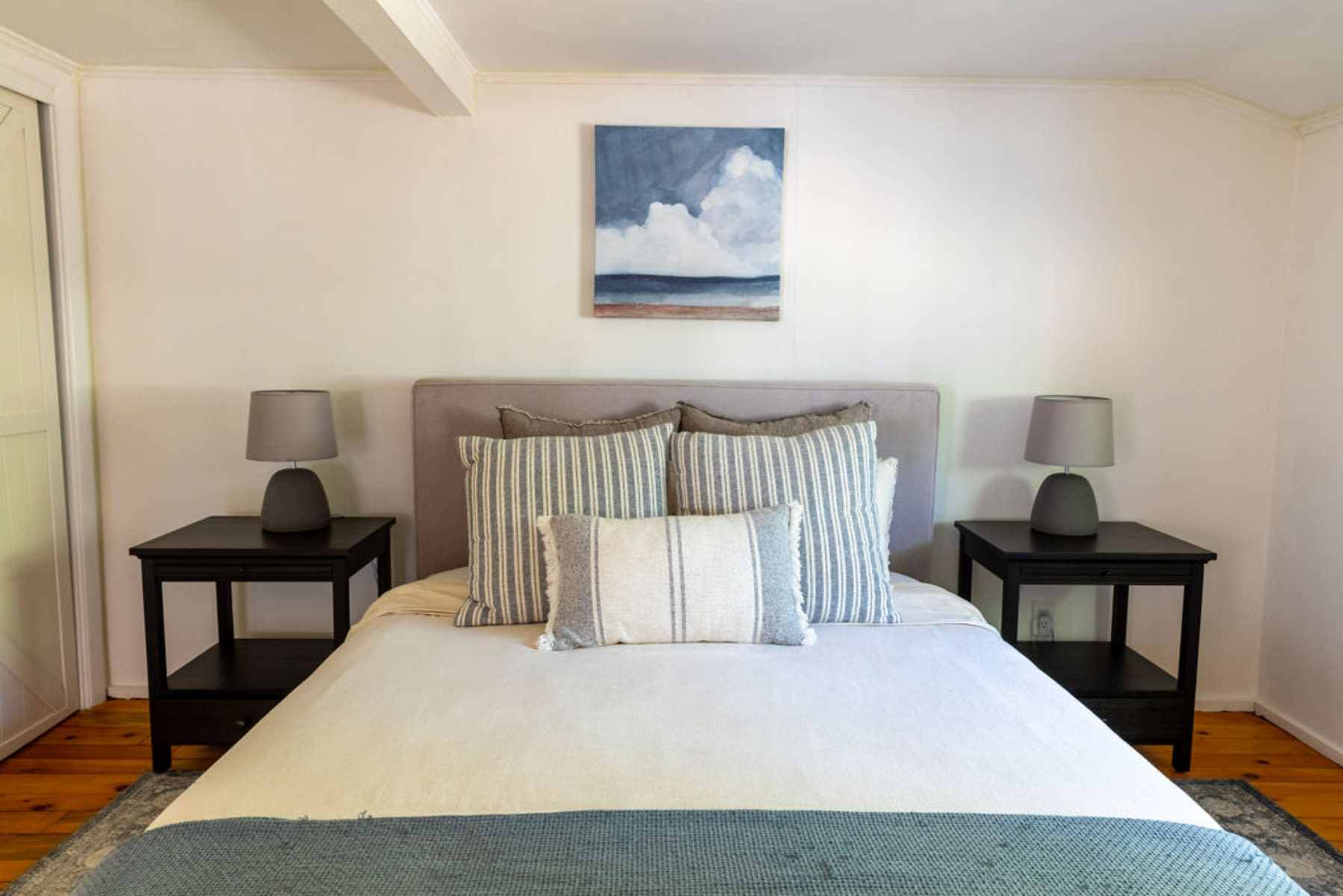 Serene bedroom featuring a queen bed with a grey upholstered headboard, flanked by black nightstands and matching grey lamps. A contemporary abstract painting hangs above the bed, complementing the cool-toned bedding.