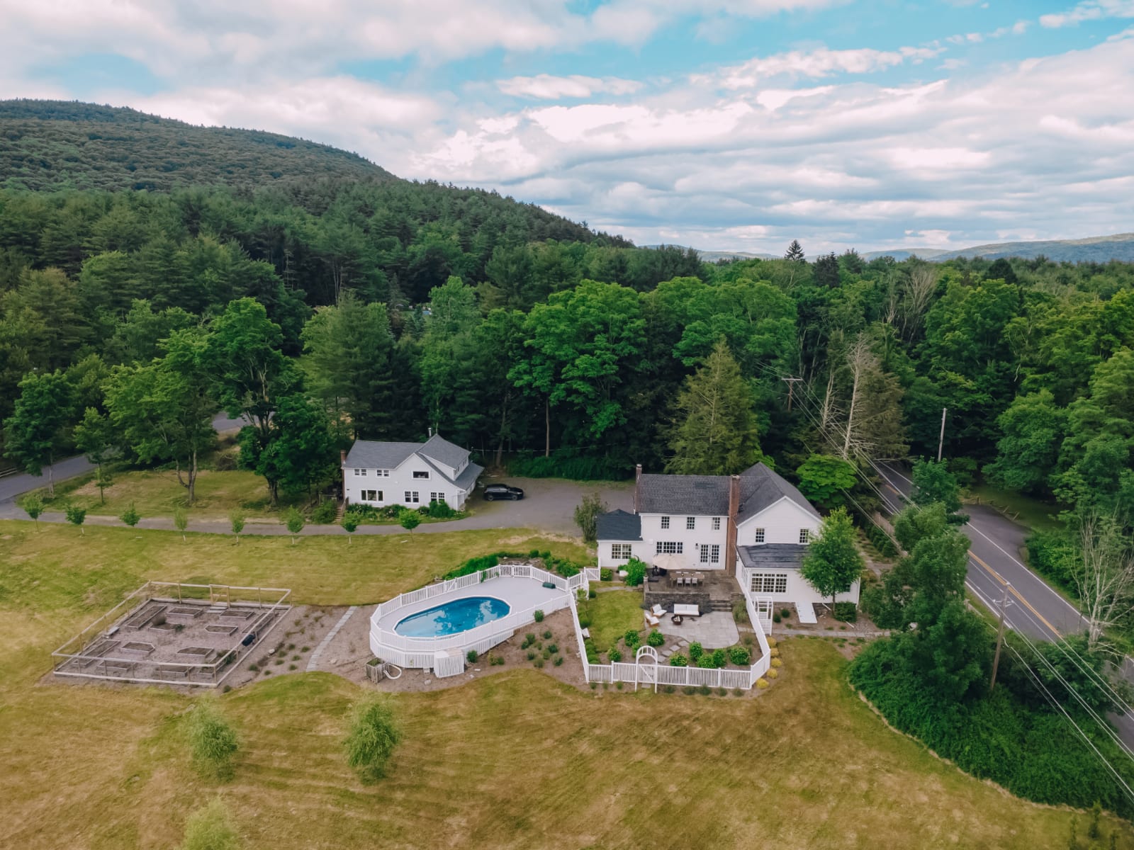 Expansive aerial view of a white farmhouse property nestled against a forested mountain backdrop. It features a fenced pool, a separate guesthouse, and a large garden plot, all set within a vast green landscape.