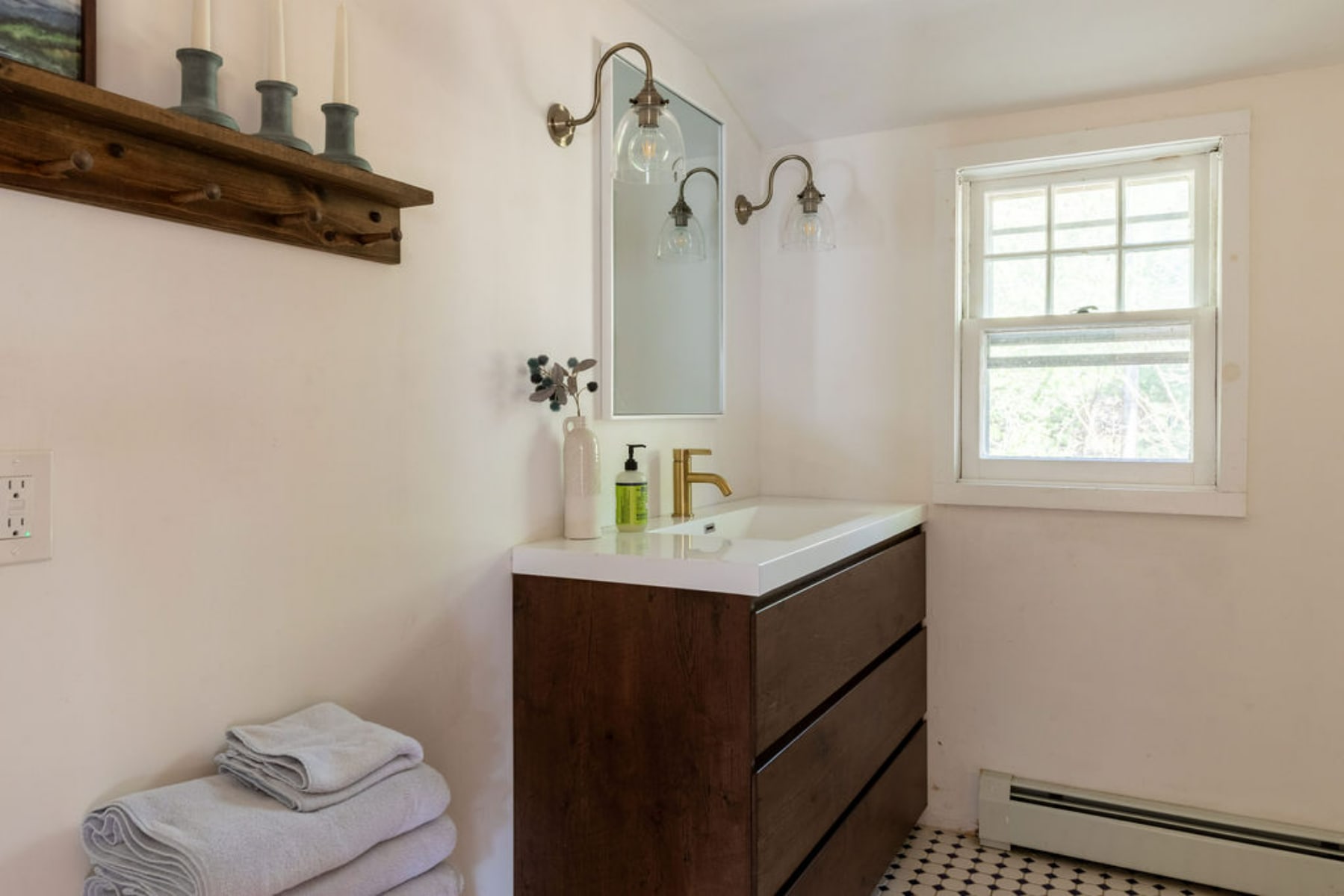 Modern bathroom vanity with a dark wood cabinet, white countertop, and gold-toned faucet. A rectangular mirror is flanked by two sconces, and a stack of fresh towels sits beside the vanity.