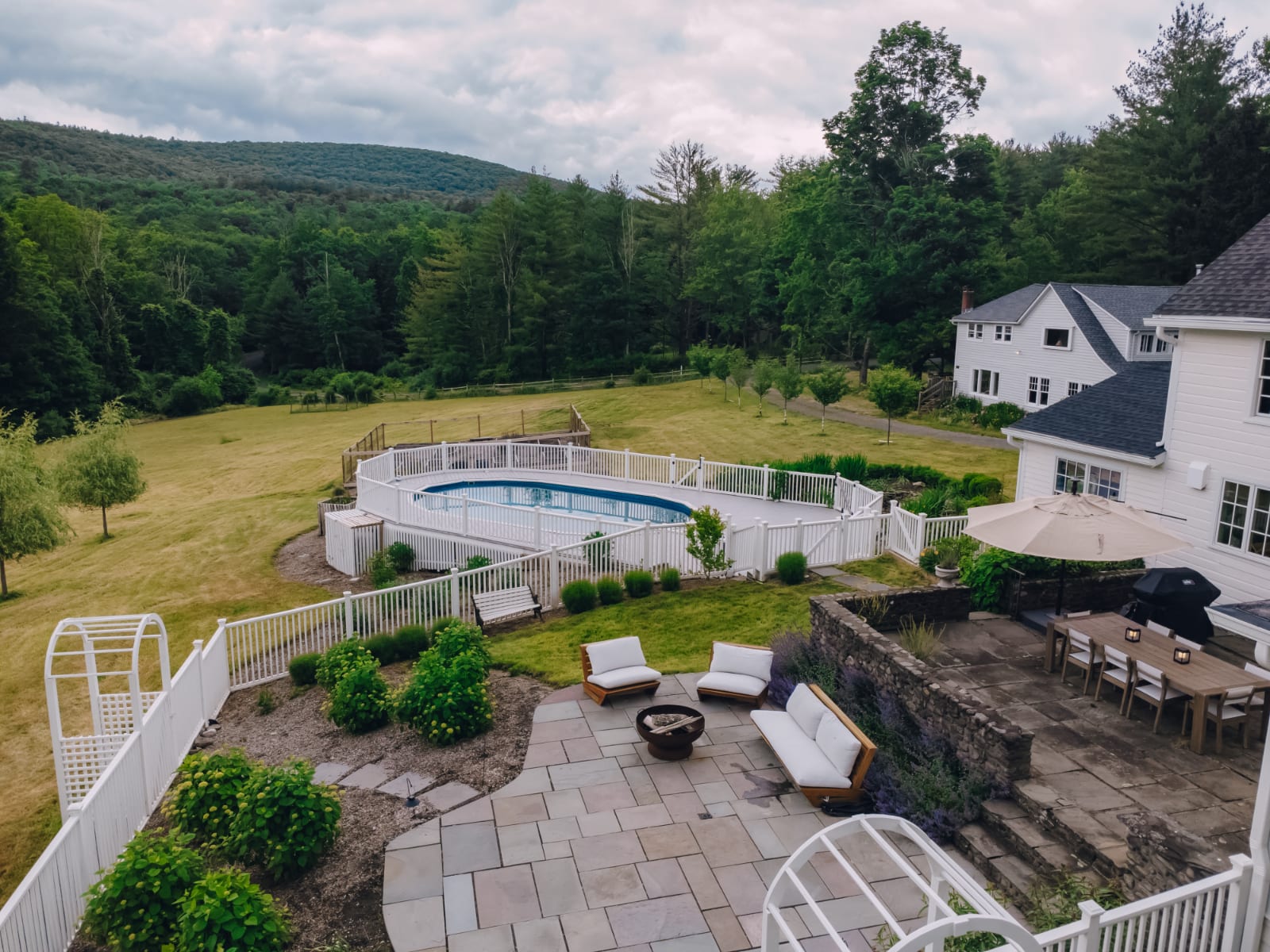 Elevated view of a white farmhouse property featuring a large inground pool surrounded by a white fence, a stone patio with outdoor seating and dining, and expansive green lawns leading to a forested mountain.