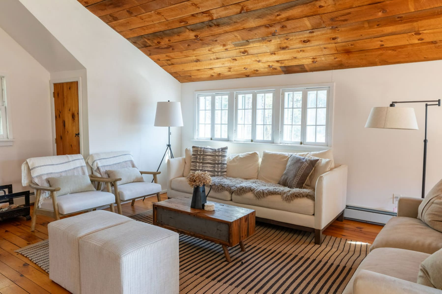 A spacious living room featuring a vaulted wooden plank ceiling, light-colored sofas and armchairs, and a striped area rug. Multiple windows provide ample natural light, and a rustic coffee table anchors the comfortable seating arrangement.