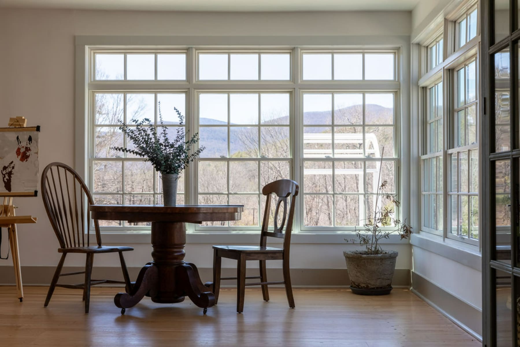 Sunlit dining area with a round pedestal dining table and two wooden Windsor-style chairs, positioned in front of a large multi-pane window offering views of distant mountains and trees. A potted plant sits in the corner.