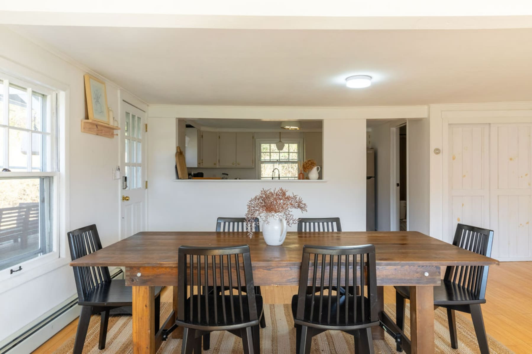 A rustic wooden dining table with six black spindle-back chairs sits on a striped rug, with a pass-through window looking into the kitchen. White walls and light wood floors create a bright and inviting atmosphere.