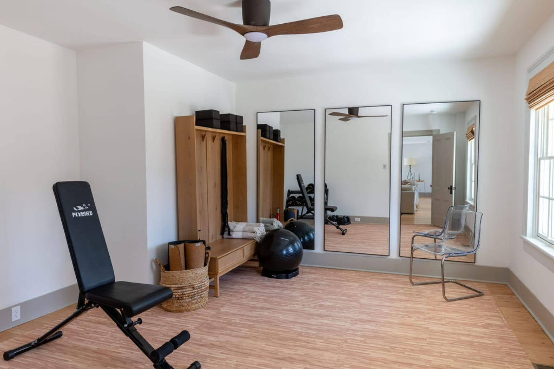 Home gym with light wood-look flooring, three large full-length mirrors against one wall, and various exercise equipment including a weight bench, yoga mats, and an exercise ball. A ceiling fan is overhead.