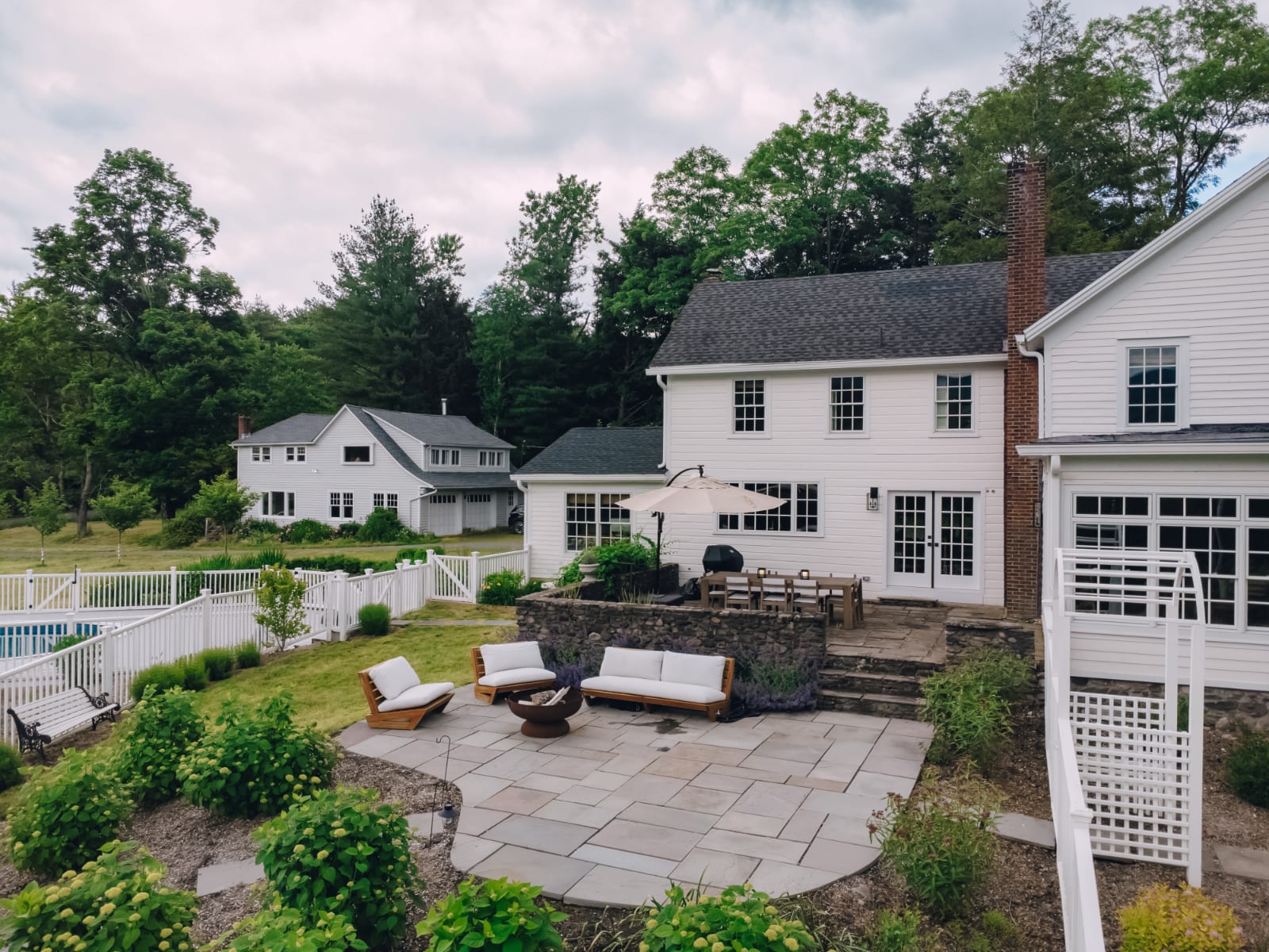 An aerial view showcases the white farmhouse with multiple outdoor living spaces, including a stone patio with dining and lounge areas, a swimming pool, and a fenced yard surrounded by lush trees.