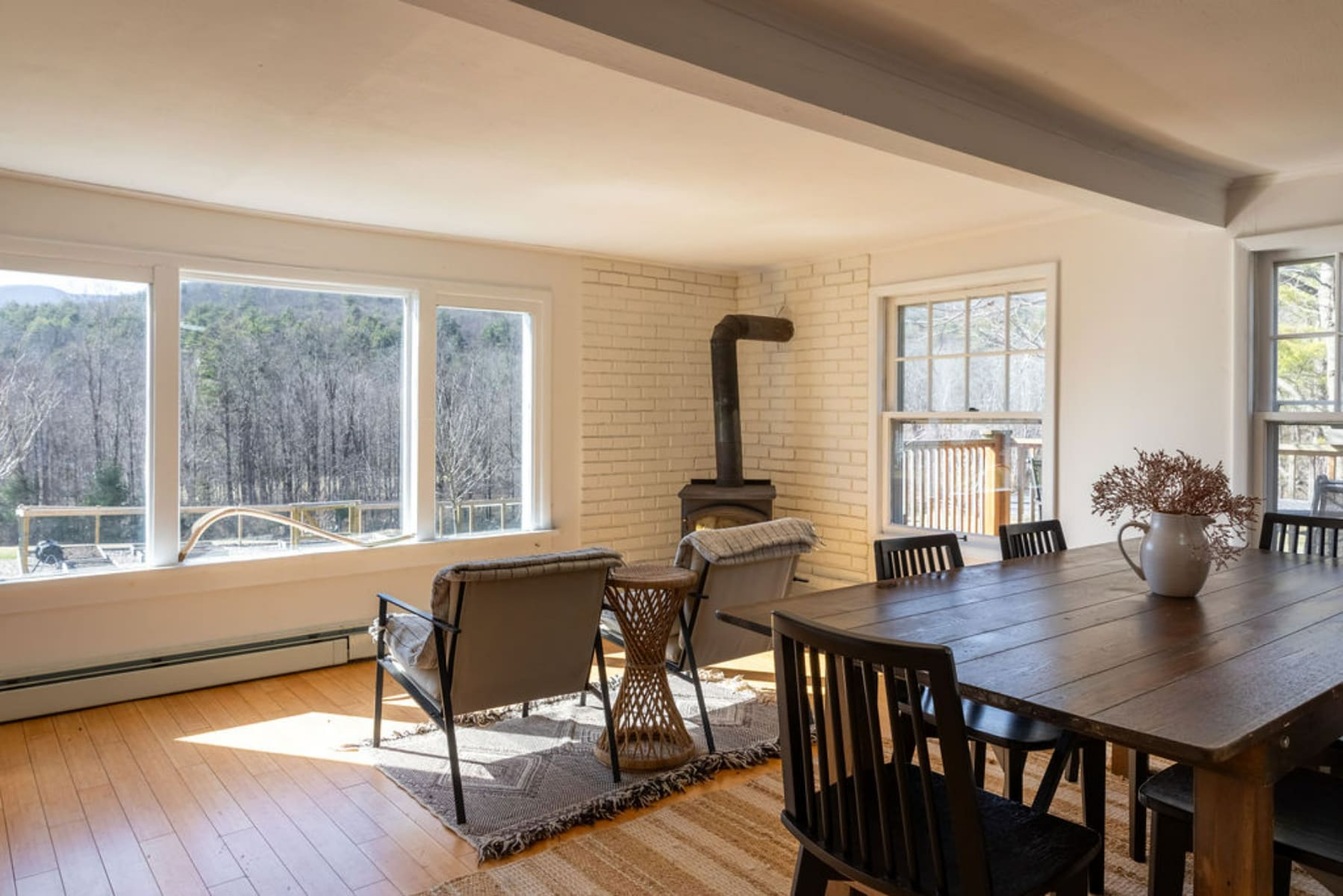 A bright living space with light wood floors, a black wood stove, and large windows offering panoramic views of a winter forest and mountains. A rustic wooden dining table and chairs are visible on the right, creating a combined living and dining area.