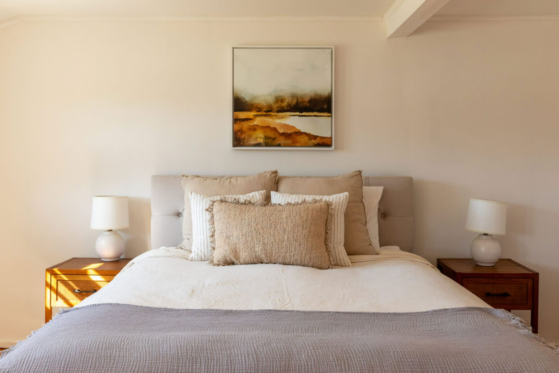 Cozy bedroom featuring a queen-sized bed with a tufted headboard, neutral bedding, and decorative pillows. Two wooden nightstands with white lamps flank the bed, beneath a framed abstract landscape painting.