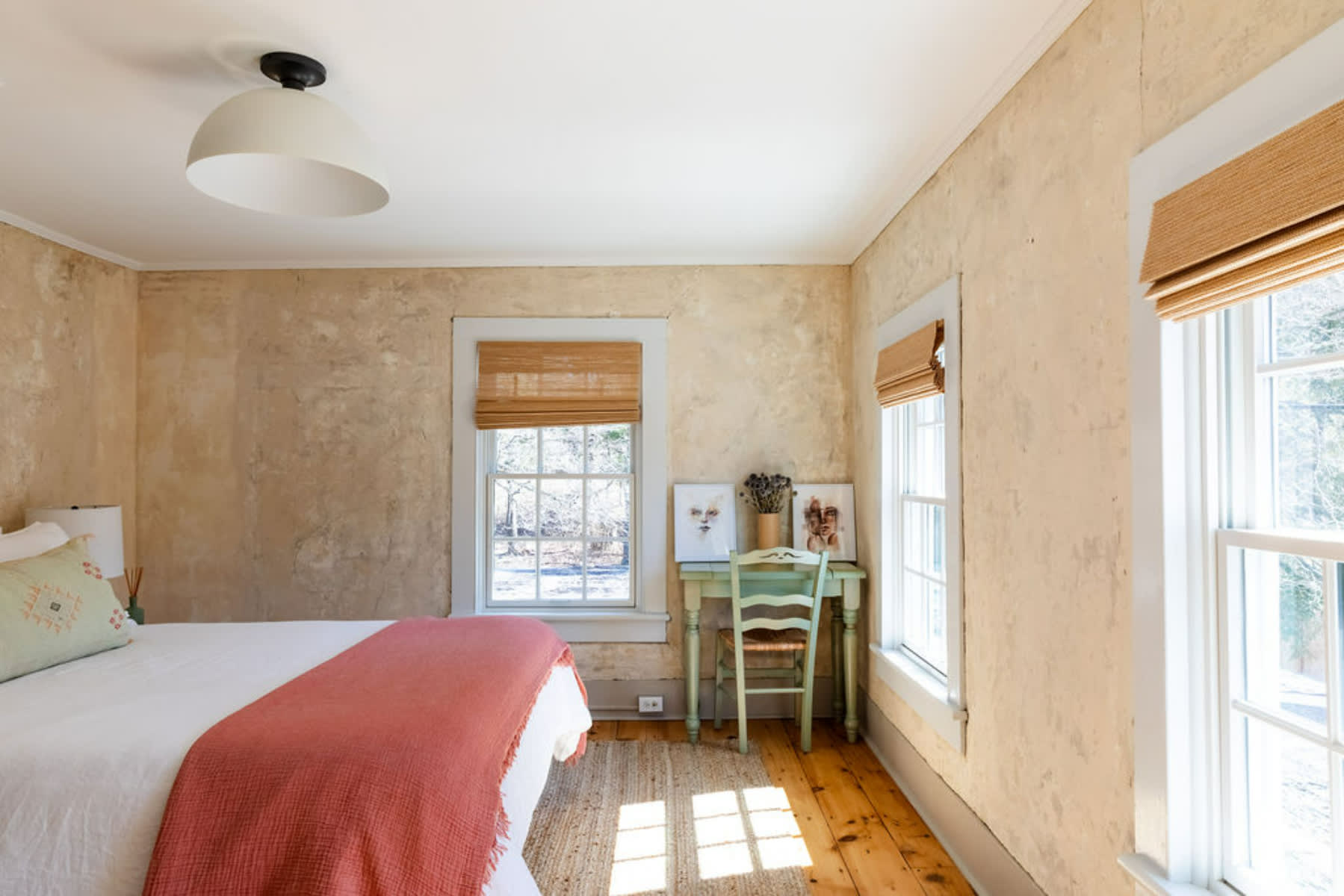 This bedroom showcases its distinctive textured walls, warm hardwood floors, and multiple windows adorned with woven blinds, alongside a small green desk and chair.