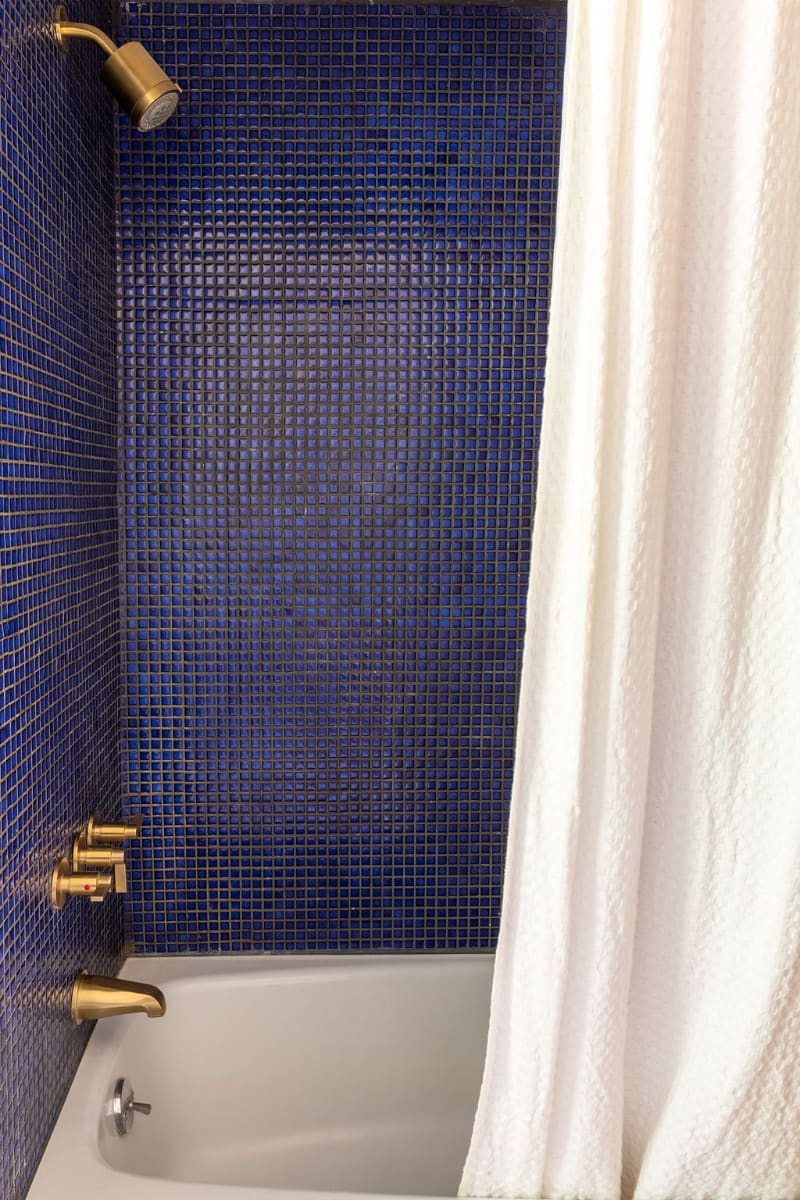 Vibrant shower and bathtub area featuring striking blue mosaic tiles covering the walls. Gold-toned fixtures, including the showerhead and faucet, add a touch of elegance against the white tub.