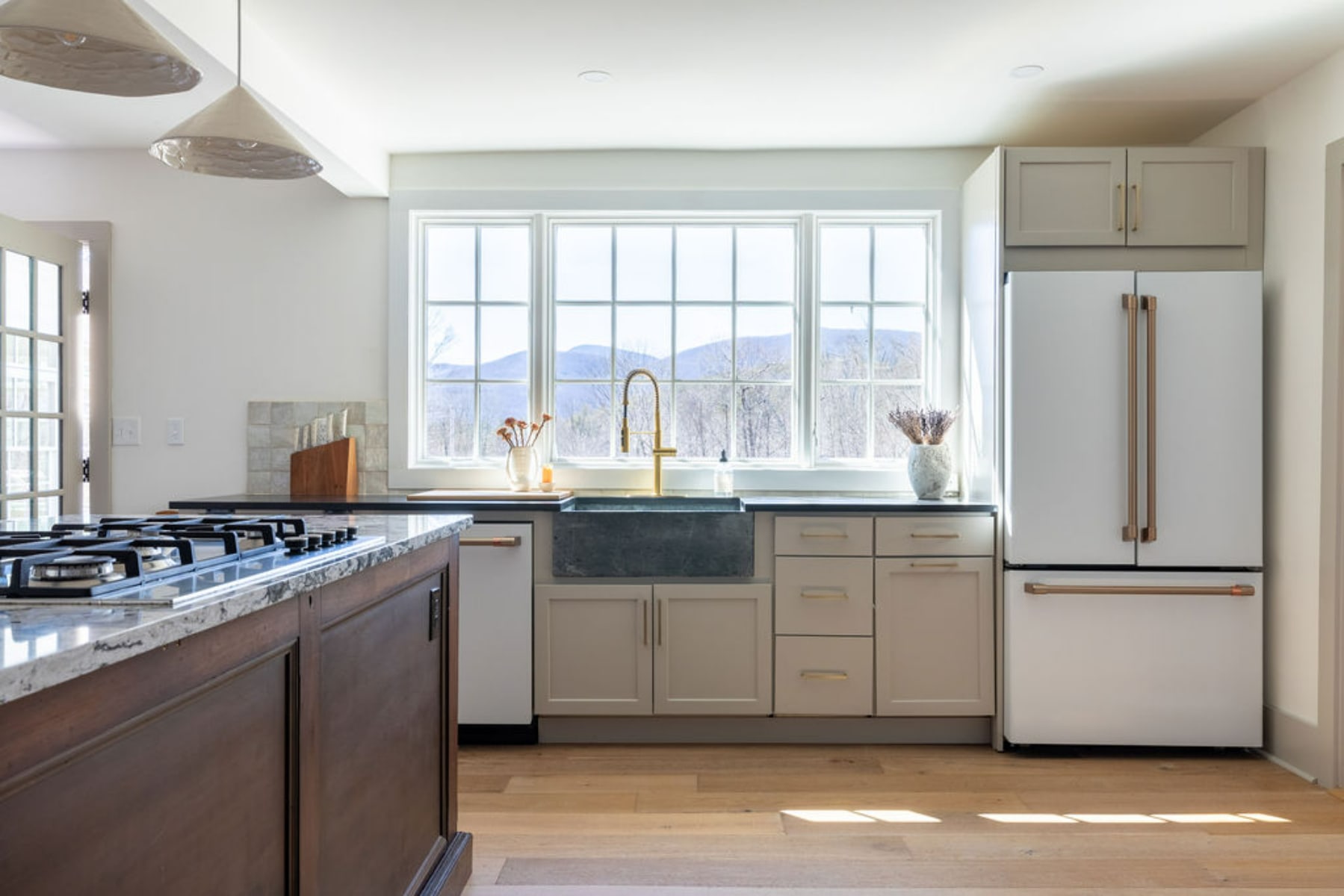 Modern kitchen with light gray cabinetry, a large window above a dark farmhouse sink with a gold faucet, and white appliances including a refrigerator. The window offers scenic views of mountains and trees.
