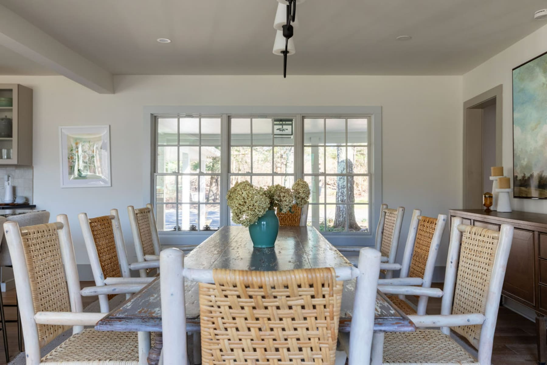 Rustic dining room with a long distressed wooden table surrounded by woven-back chairs, set in front of a large window overlooking the backyard, with recessed lighting and a modern chandelier.