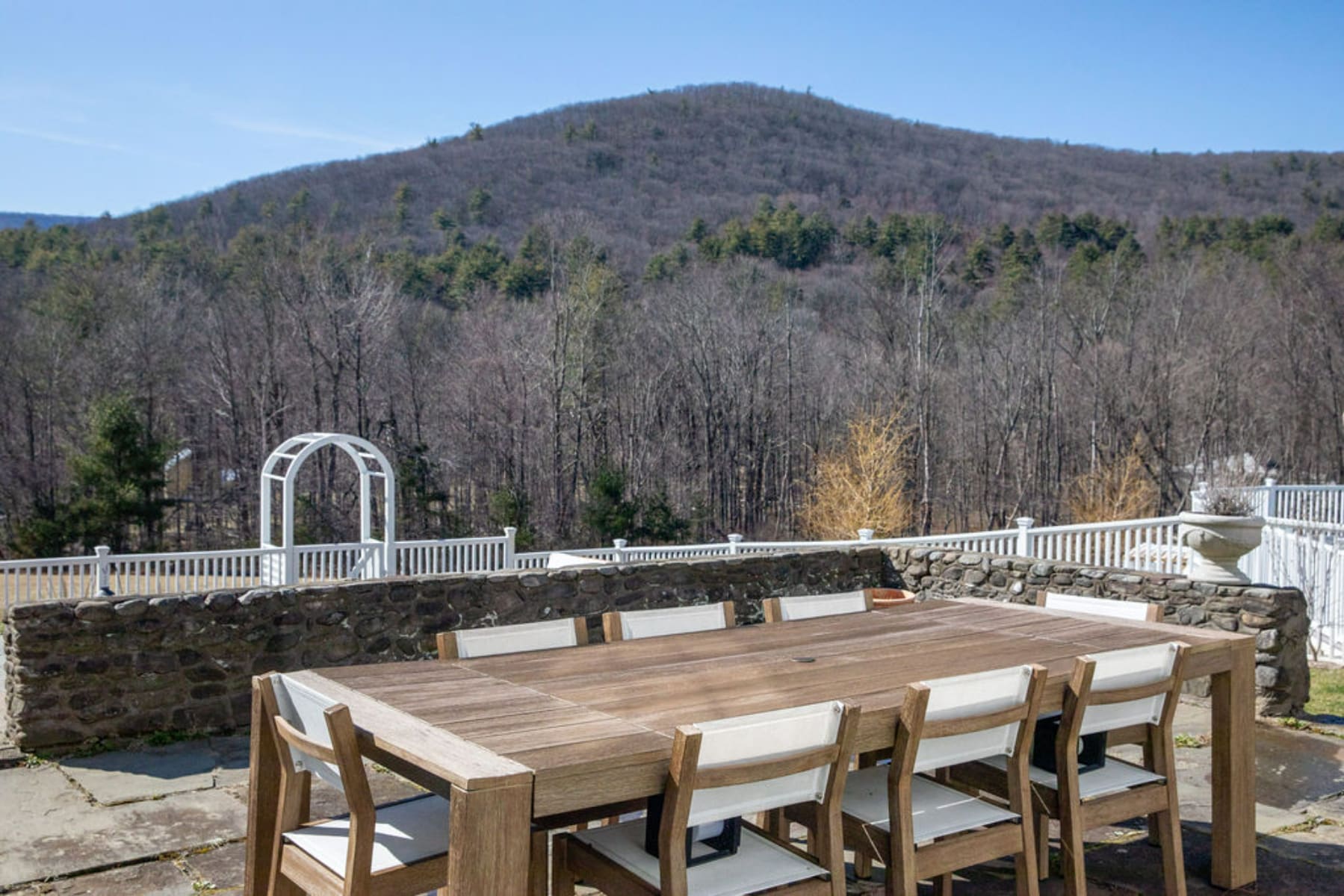 A large wooden outdoor dining table with eight chairs sits on a stone patio, offering expansive views of a forested mountain range under a clear blue sky. A white railing and stone wall define the patio's edge.